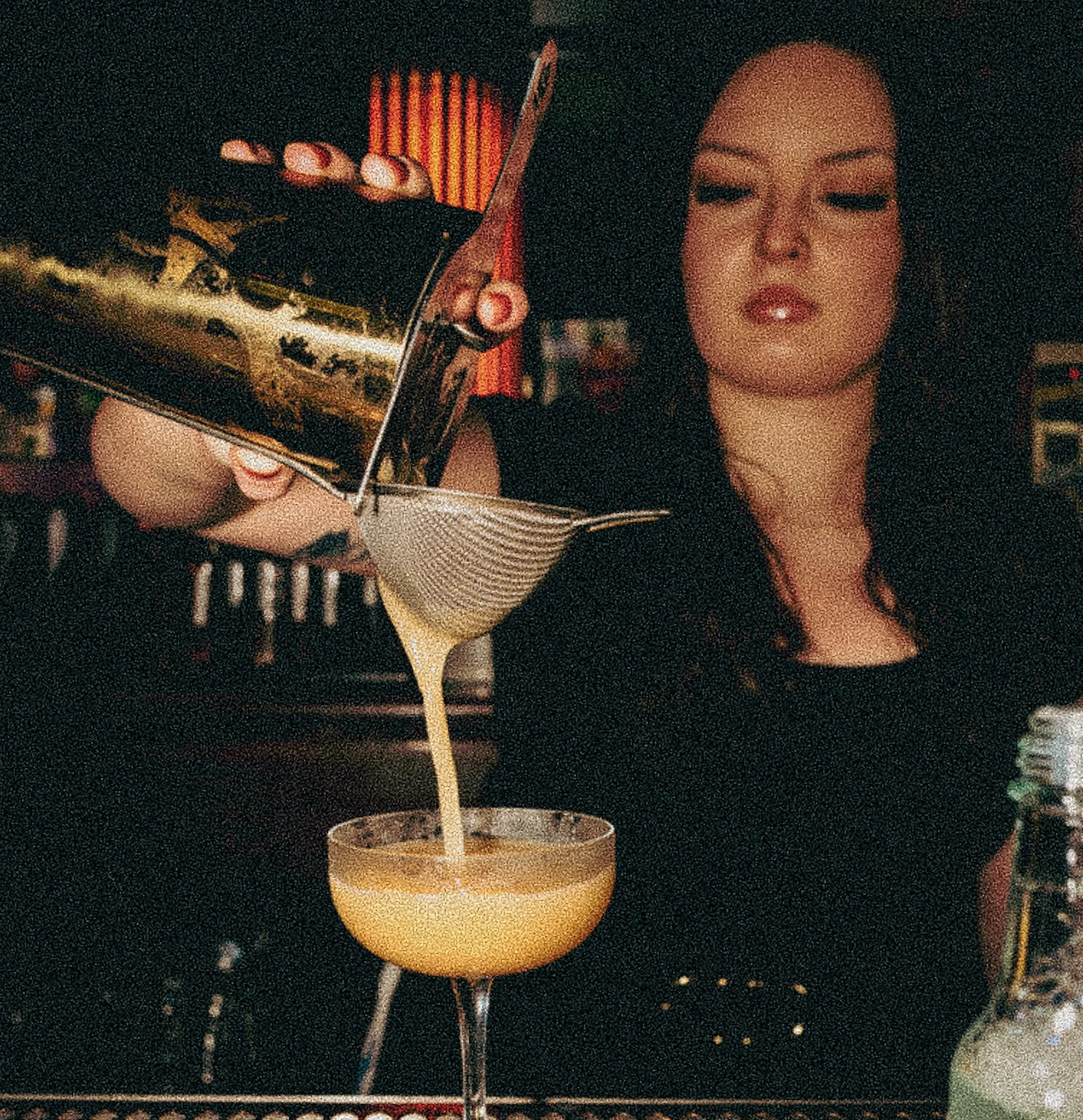 A woman pours a yellow cocktail through a cocktail strainer into a glass.