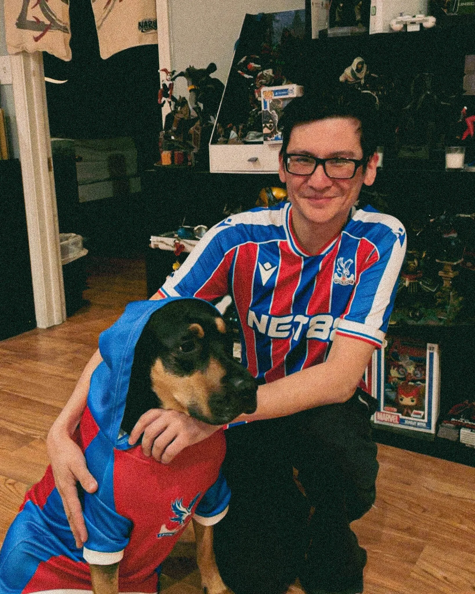 A young man with glasses smiling while kneeling next to a dog dressed in a sports jersey and cape. The background shows a room with shelves and various collectibles.