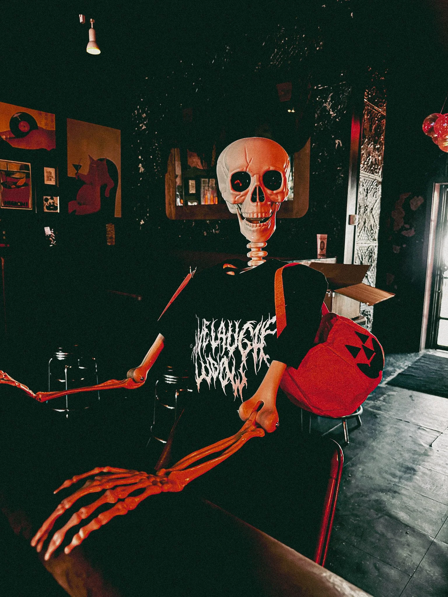Skeleton with a human skull mask sitting at a bar, wearing a black t-shirt with white text, in a dimly lit room with Halloween decorations.