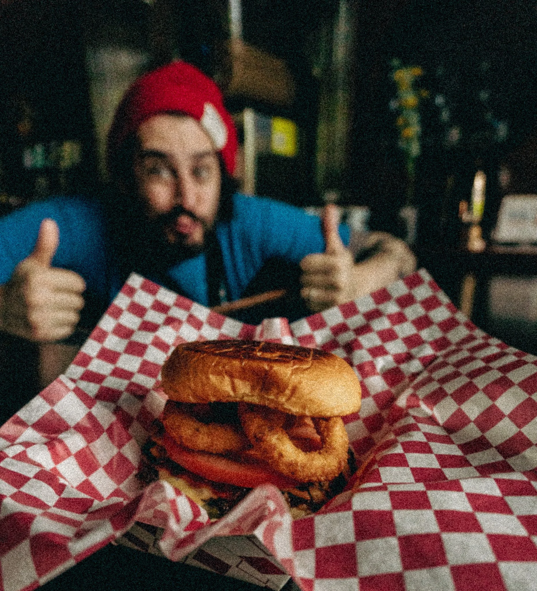 A man with a beard, wearing a red cap and blue shirt, giving two thumbs up behind a burger with fried onion rings, tomato, lettuce, and bun on checkered paper.