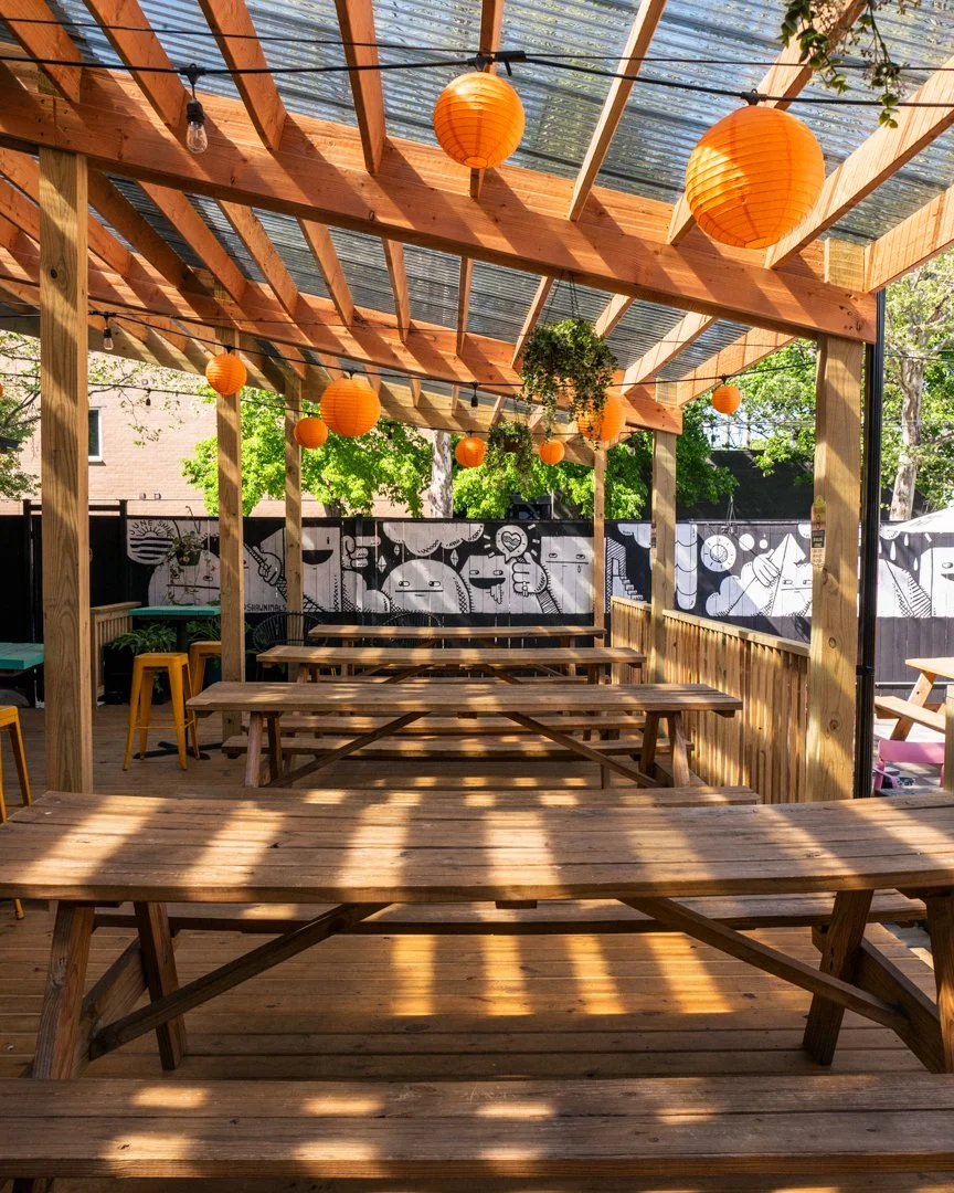An outdoor patio behind Ludlow Lliquors, a neighborhood bar,  in the Avondale and Logan square area of Chicago. Spacious seating with large picnic tables and hanging string lights with orange lanterns.