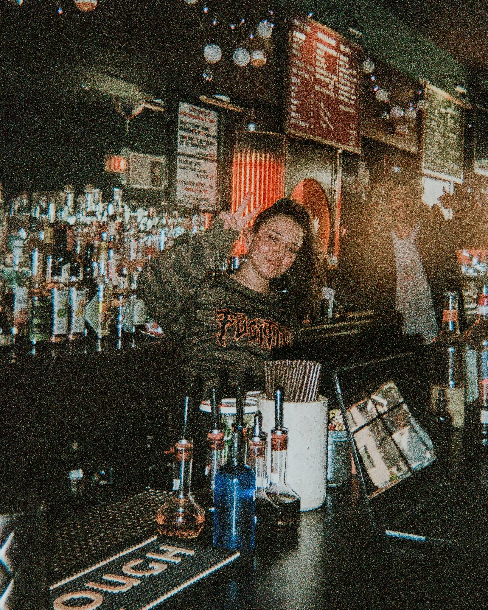 A woman smiling behind a bar counter in a dimly lit bar, with bottles and bar tools in the foreground and a menu board on the wall in the background.