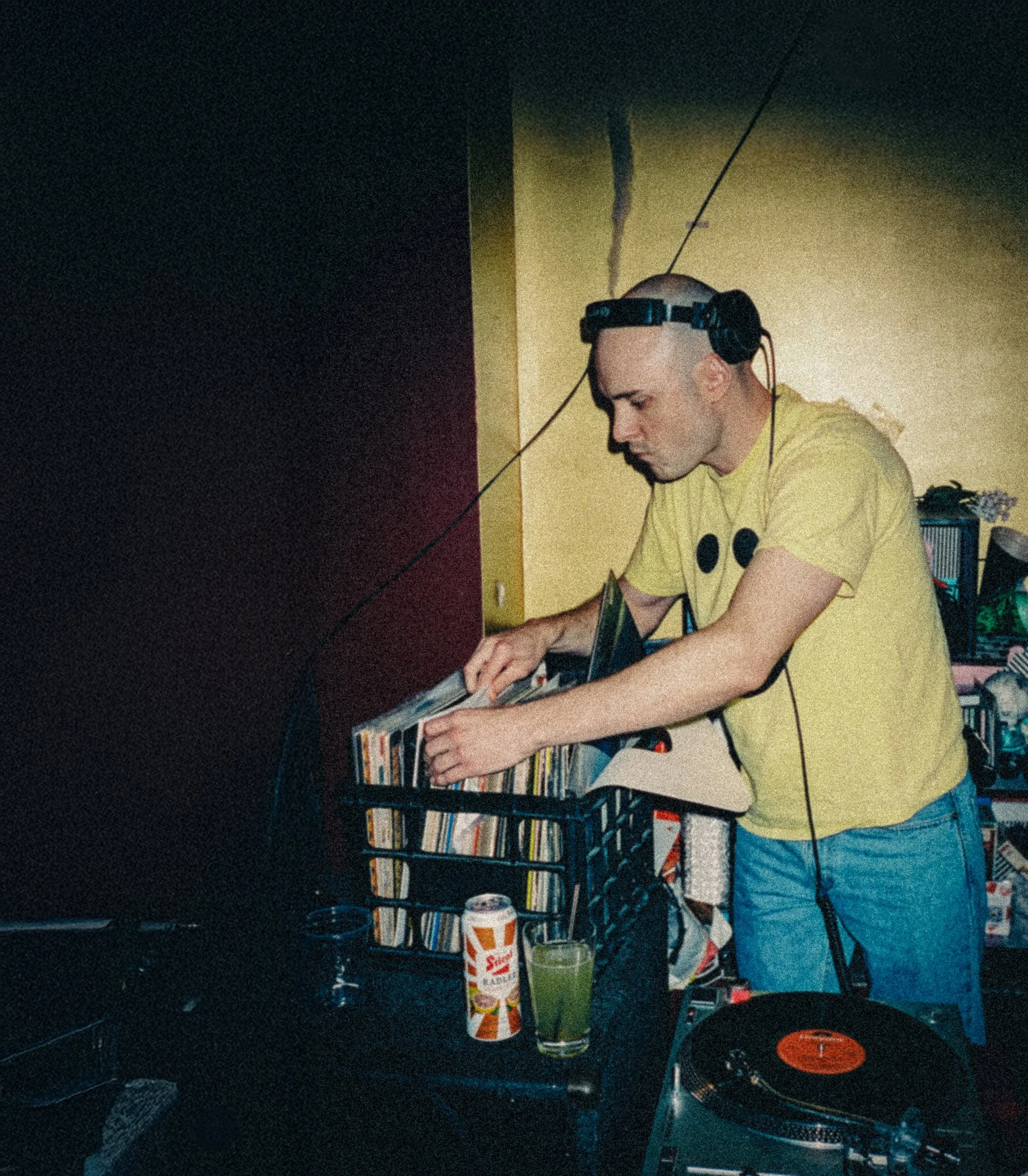 A man wearing a yellow t-shirt, blue jeans, and headphones is DJing with vinyl records at a party. There is a beverage and a can on the table, and shelves filled with vinyl records in the background.