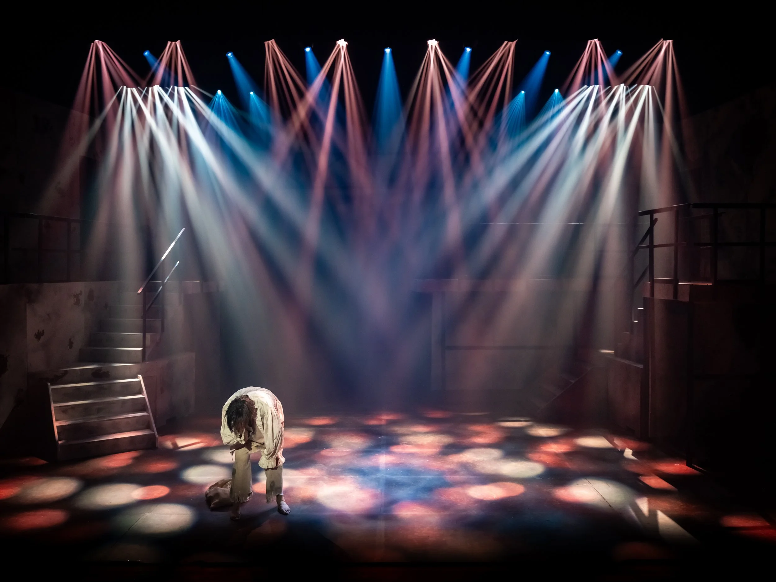 A person bowing on stage with colorful lights shining down, stairs on either side, and shadows on the floor.