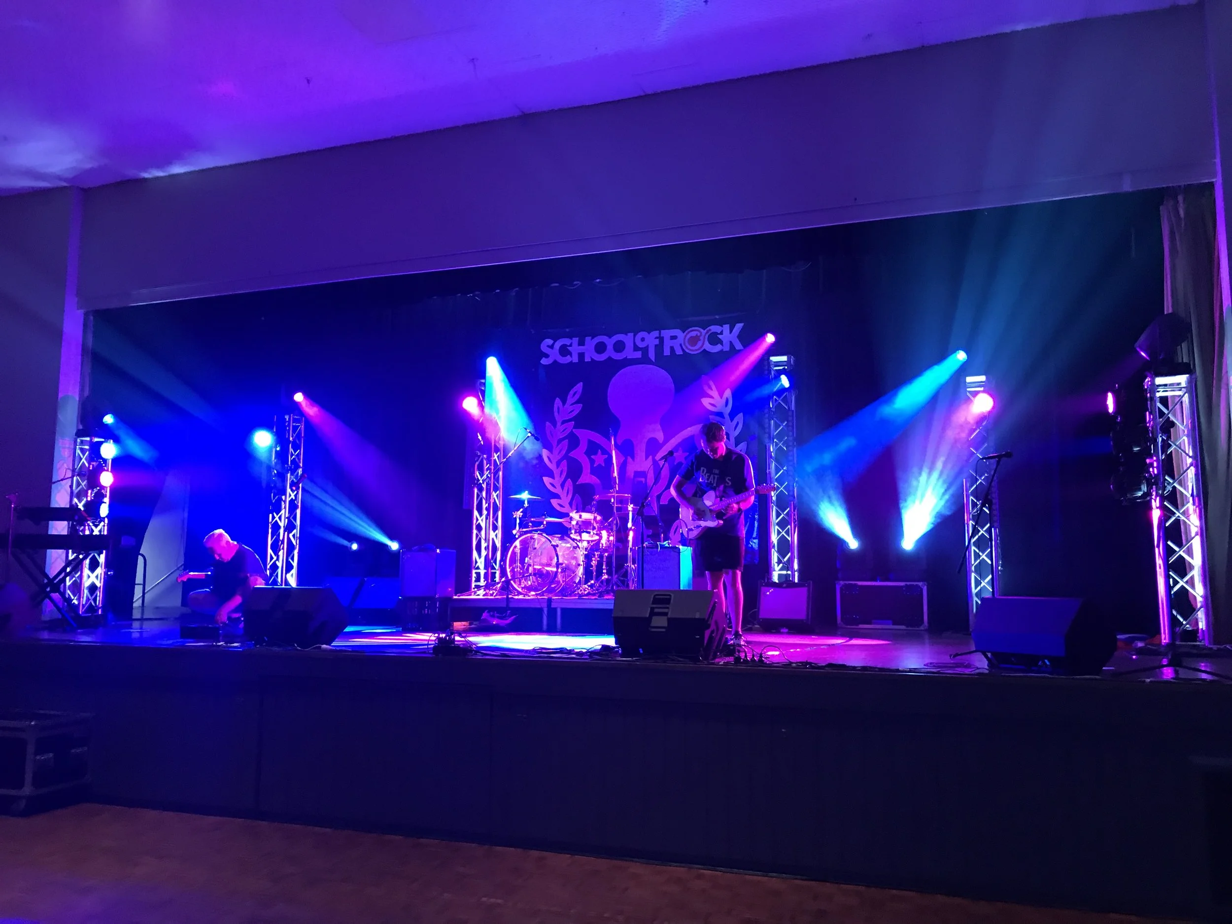 A band performs on stage with colorful purple and blue lights, with a backdrop that reads 'School of Rock.'