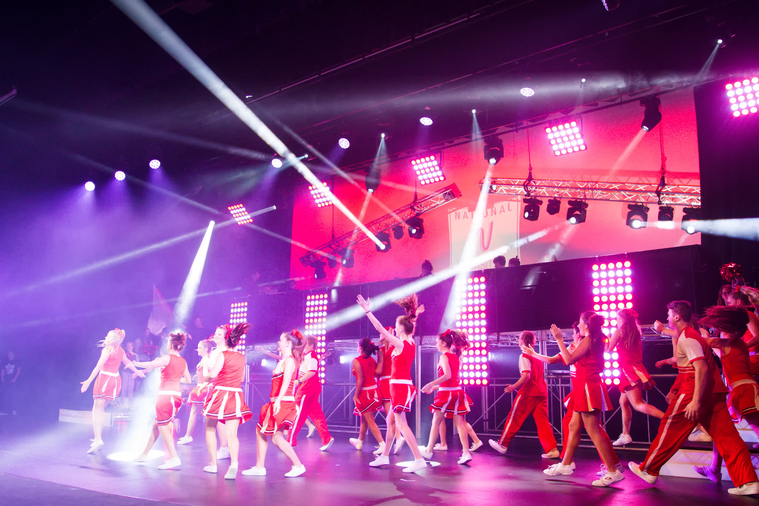 Group of performers in red costumes dancing on stage with colorful lighting, large LED screens, and bright spotlights.