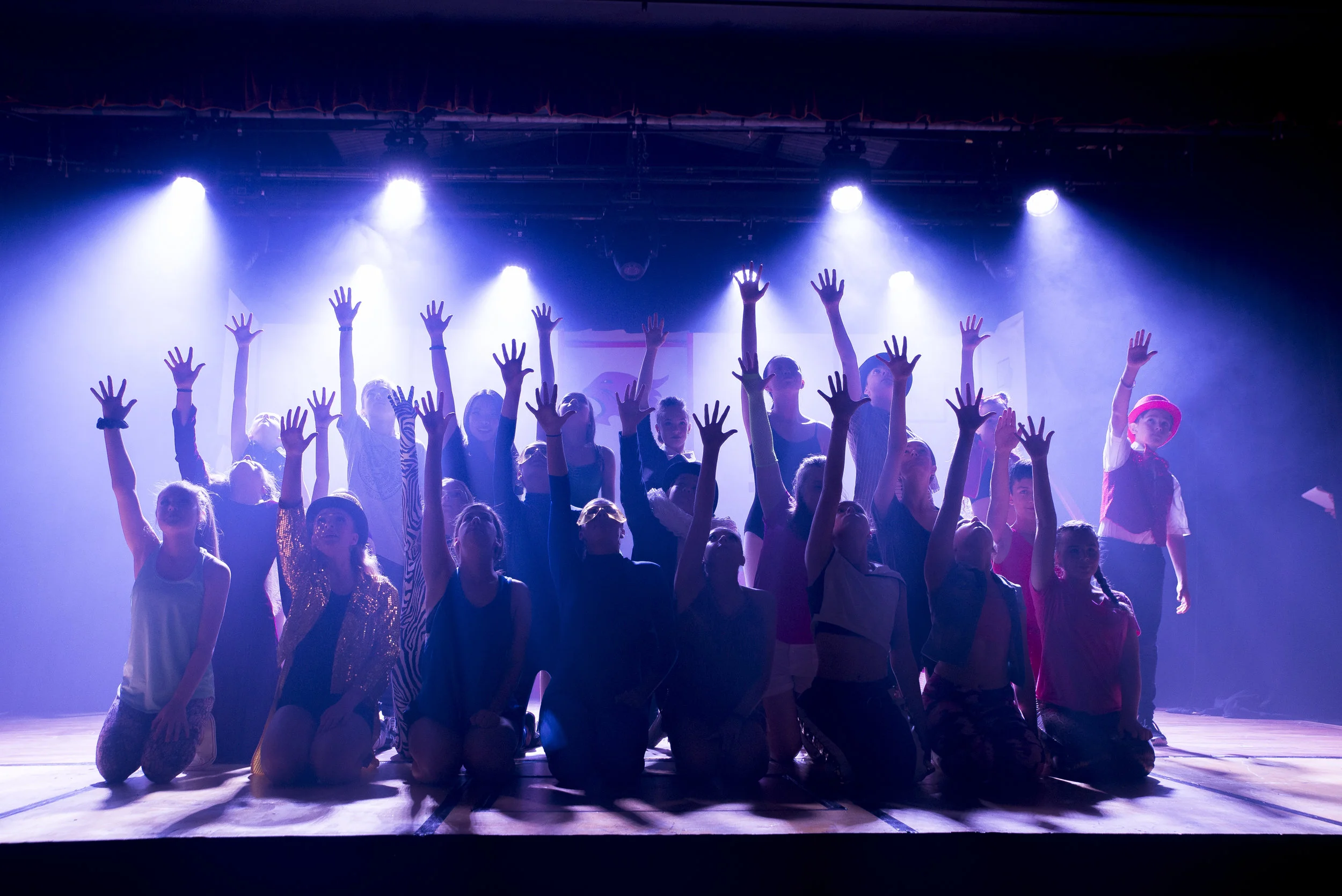 Group of children on stage performing with hands raised, stage lights overhead, in a theater or auditorium.