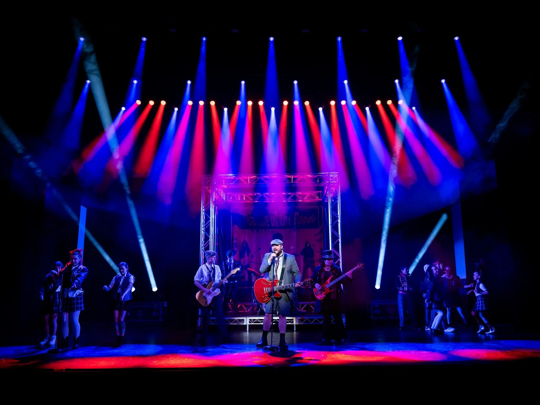 A live stage performance with a band of musicians and dancers, colorful stage lighting, and a male singer playing an acoustic guitar.