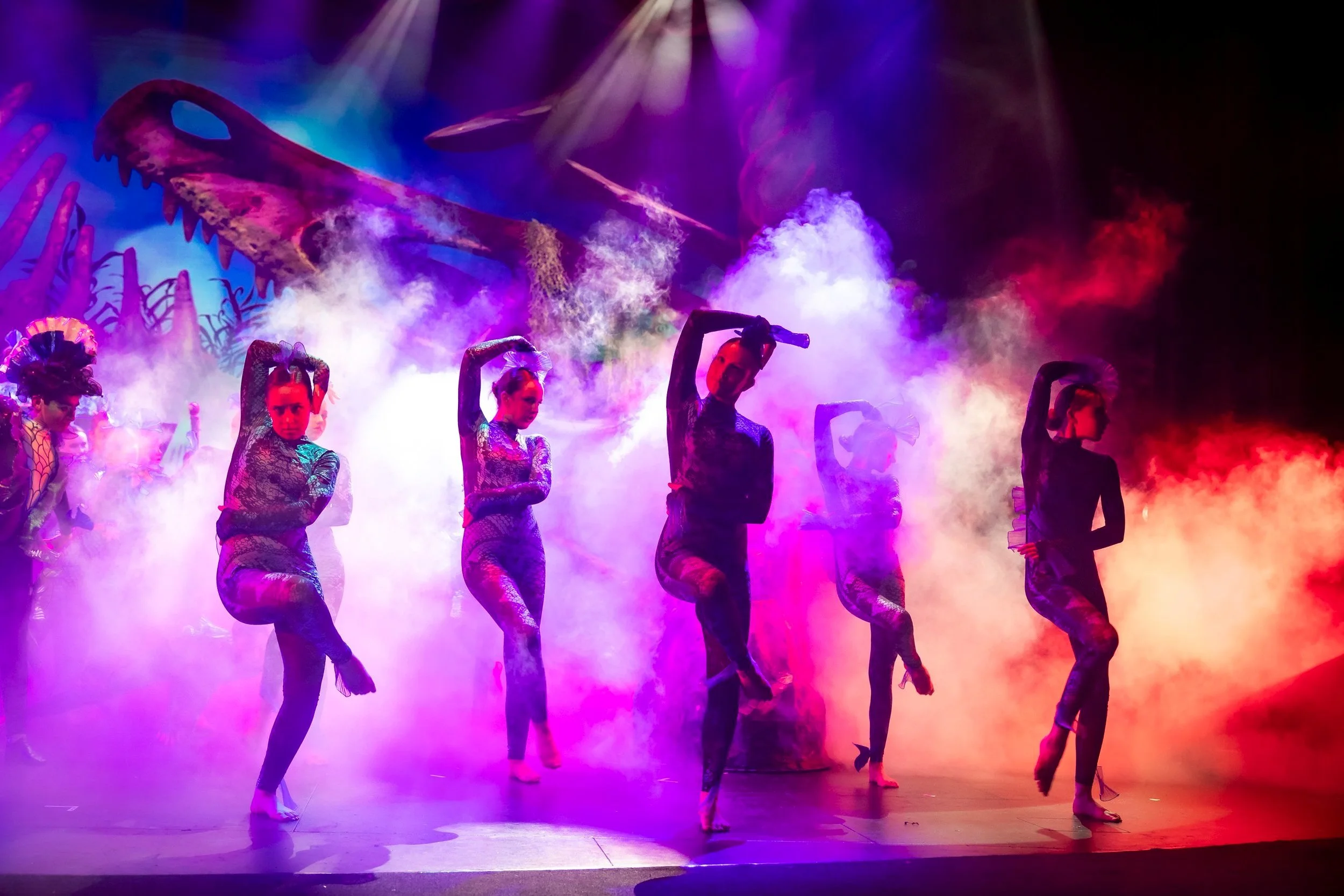 Dancers performing on stage with colorful smoky lighting and a prehistoric dinosaur backdrop.
