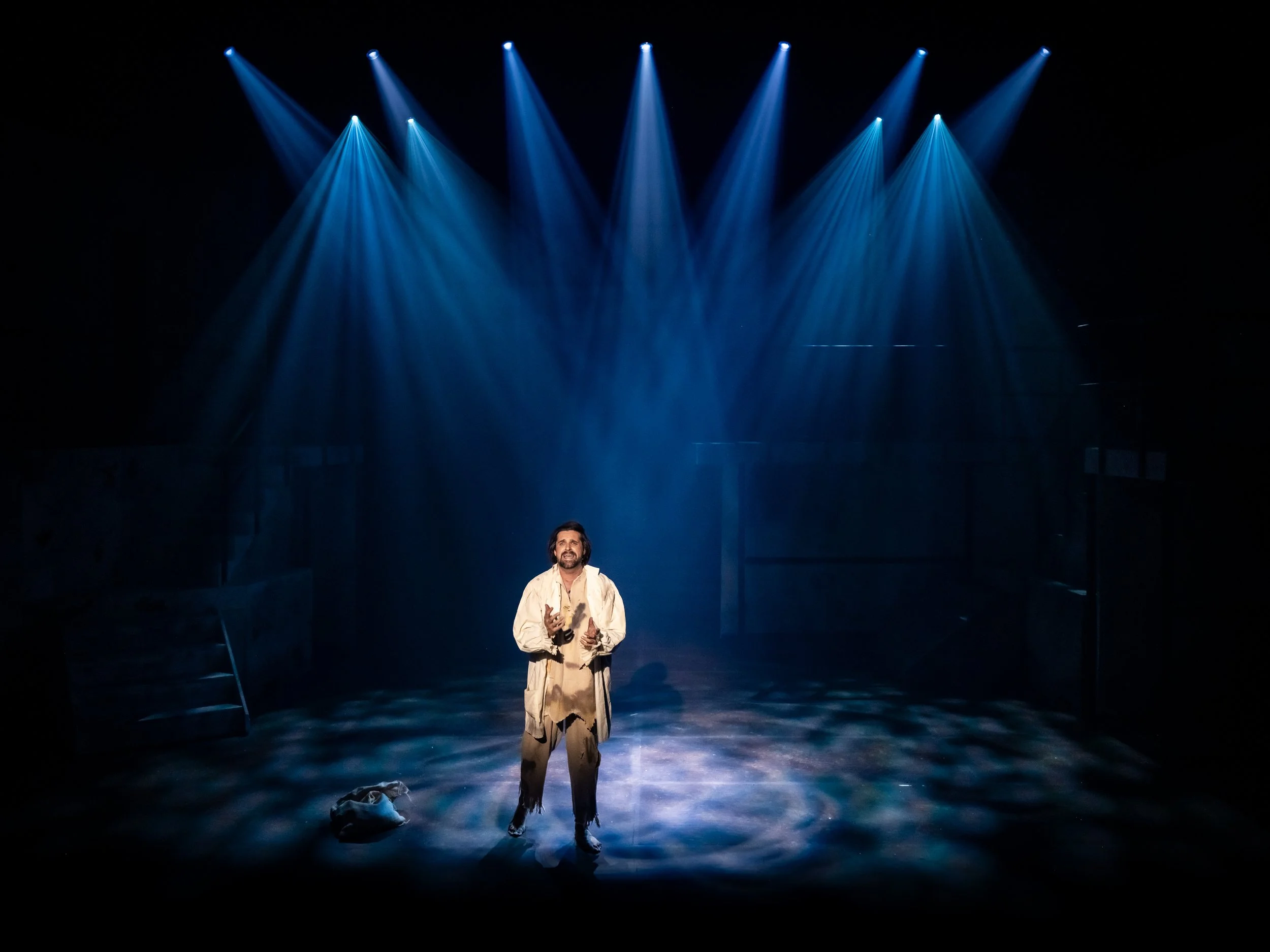 A man stands on stage under blue spotlight beams in a dark theater, wearing a beige costume with ripped pants and holding a plastic bottle, with a pair of shoes and a bag on the floor nearby.