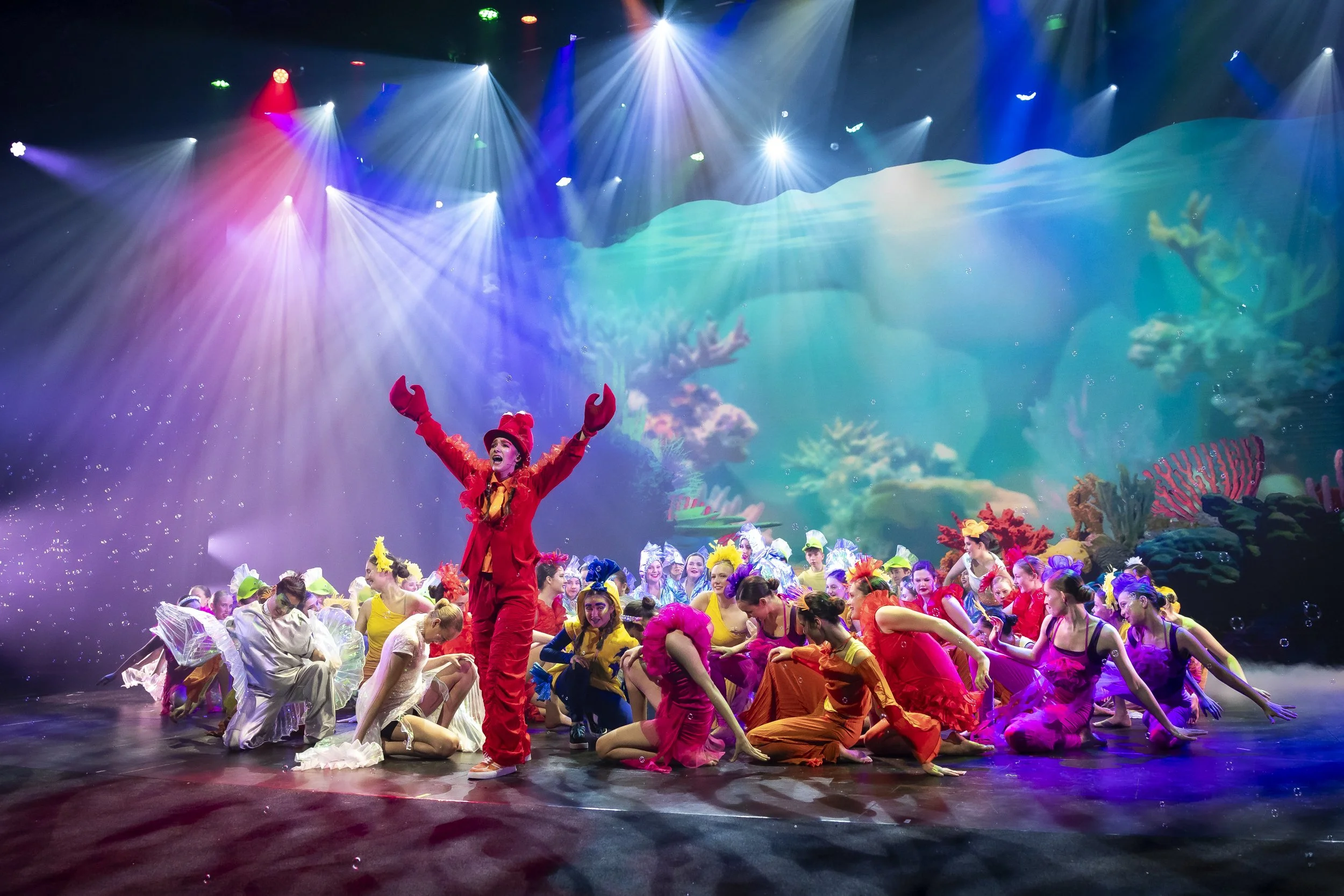 A theatrical performance on stage with a large group of performers dressed in colorful costumes. The background features a vibrant underwater scene with coral and marine life, illuminated by multicolored stage lights. A woman in red with large red gloves is standing with arms raised at the center.