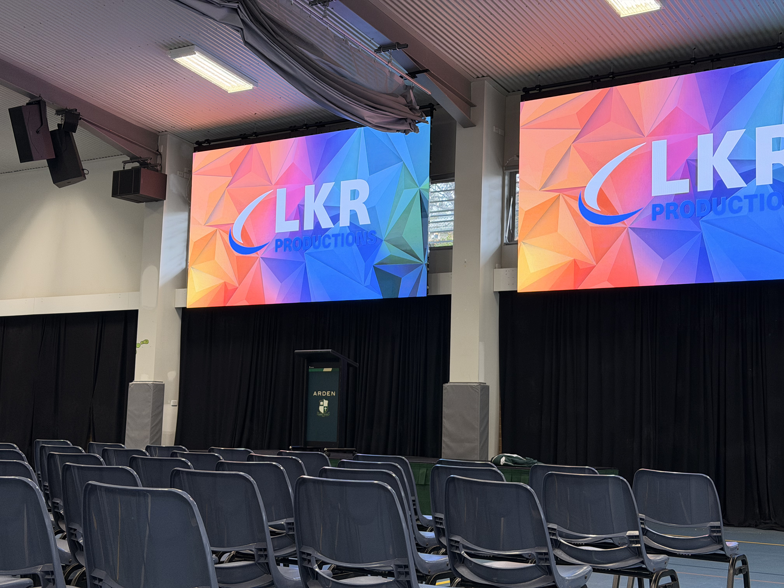 A large indoor auditorium with multiple rows of blue chairs facing a stage with two large digital screens displaying the logo of LKR Productions on a colorful geometric background. The ceiling has rectangular light fixtures and mounted speakers, with some equipment or fabric partially hanging from it.