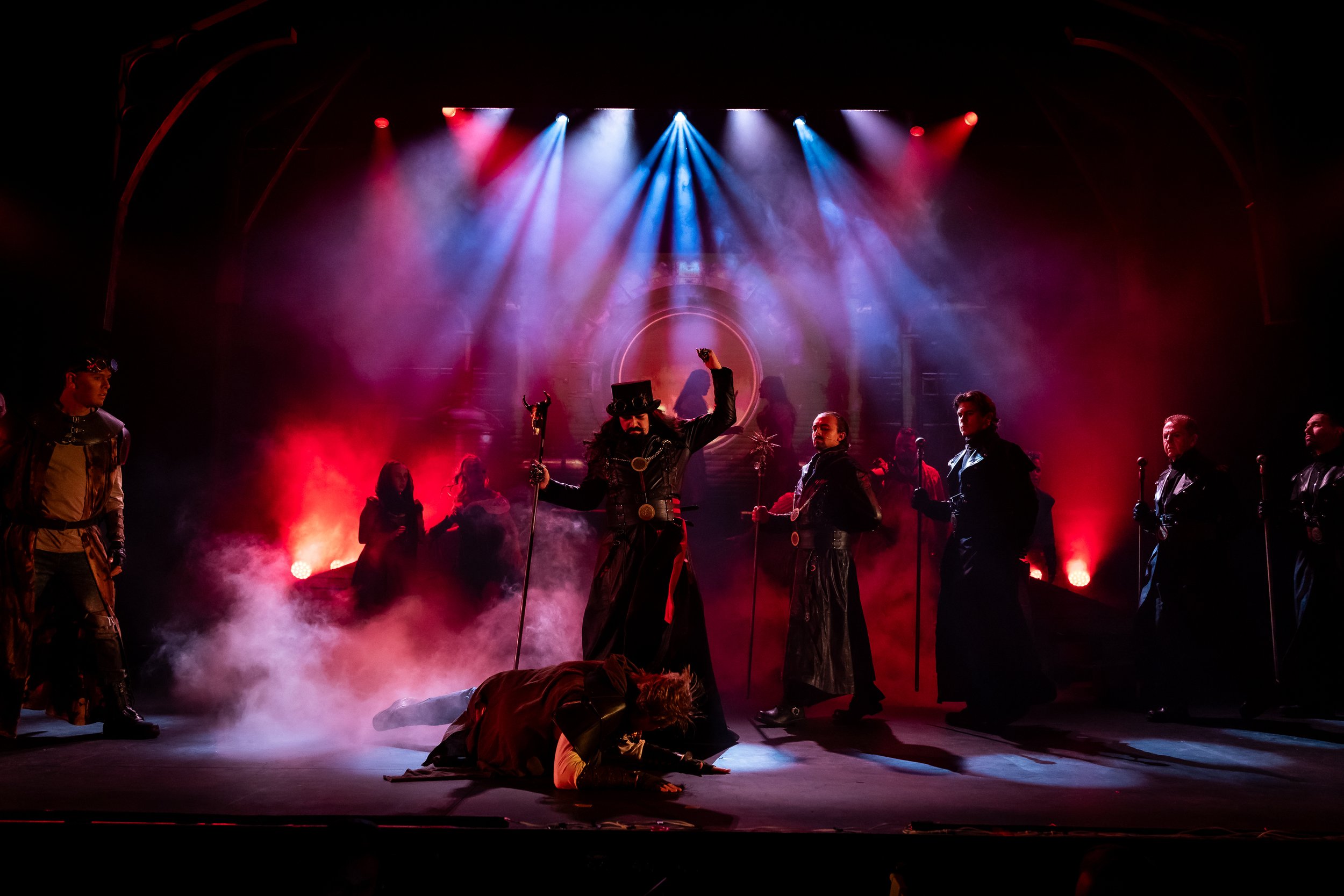 A theatrical stage performance with multiple actors in elaborate costumes, fog effects, and colorful stage lights.