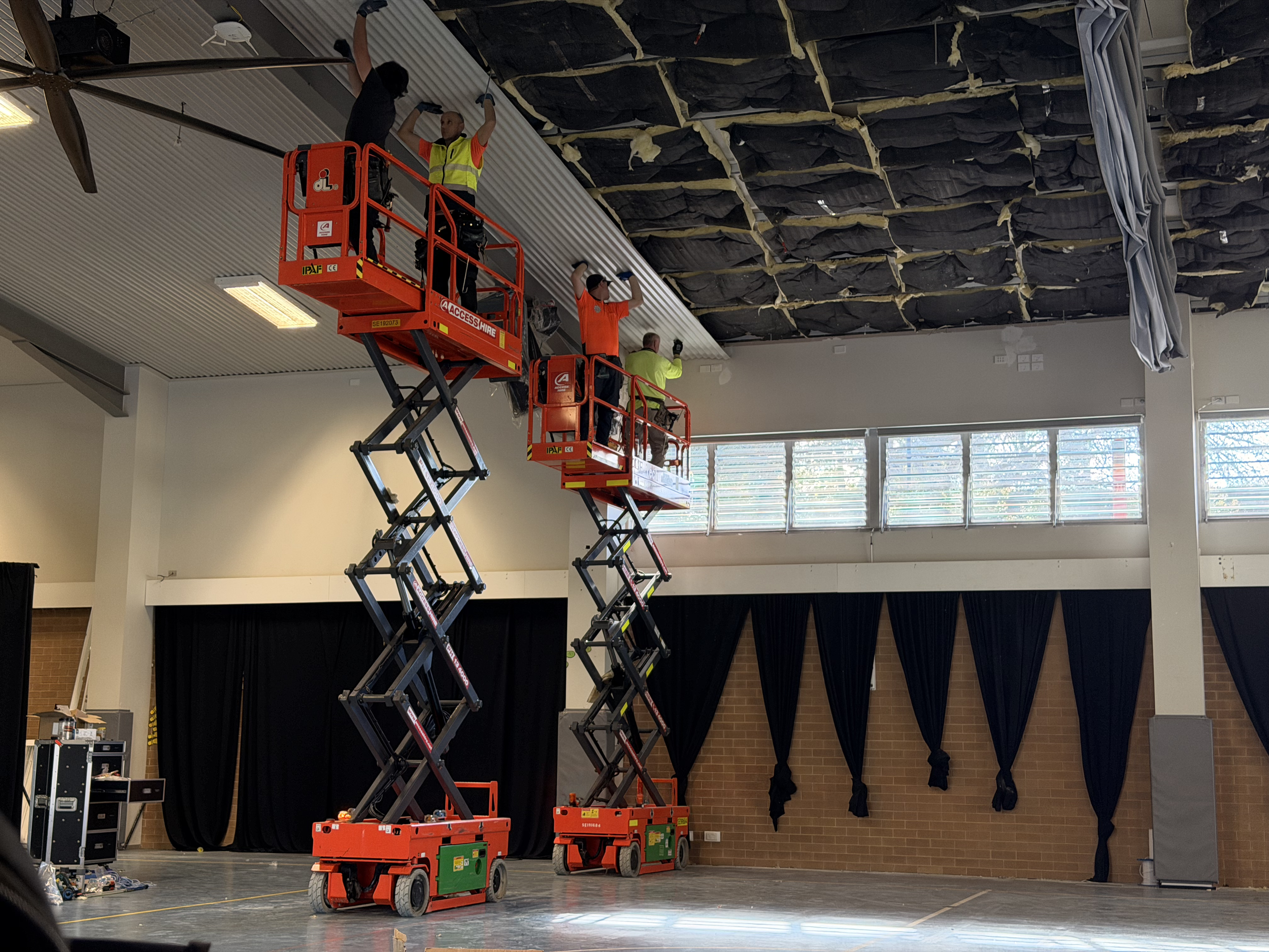 Four workers are on lift platforms installing or repairing a ceiling in a large room with black curtains and large windows.