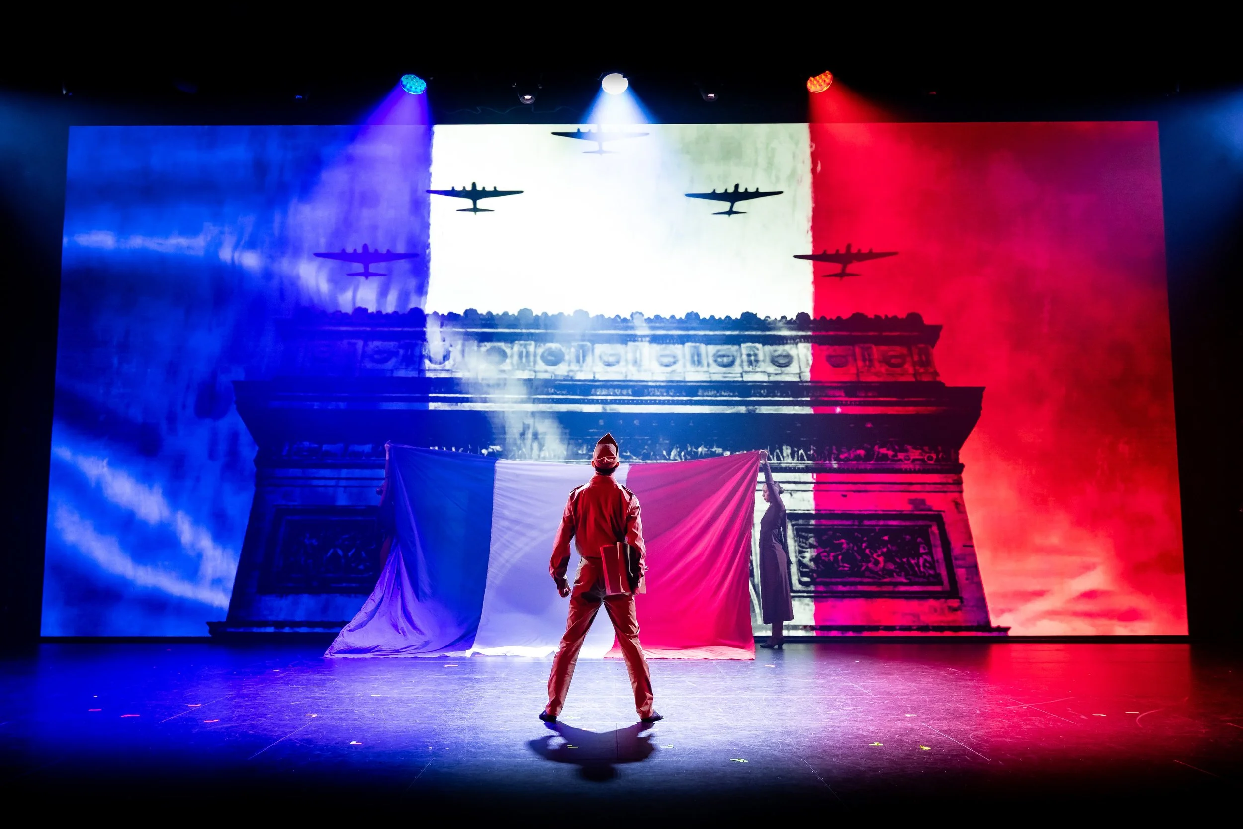 Person standing on a stage with a large backdrop featuring a silhouette of the Eiffel Tower and the colors of the French flag. The scene is illuminated by stage lights in blue, white, and red.