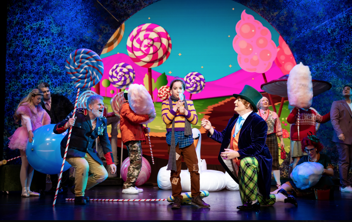 The scene is a colorful stage with a whimsical, candy-themed backdrop, featuring large, swirled lollipops and candy trees. Several people dressed in playful, eccentric costumes are present, including a girl in a purple and yellow striped sweater, a man in a blue velvet jacket and green plaid pants, and others wearing oversized candy props and bright clothing, suggesting a theatrical or performance setting.