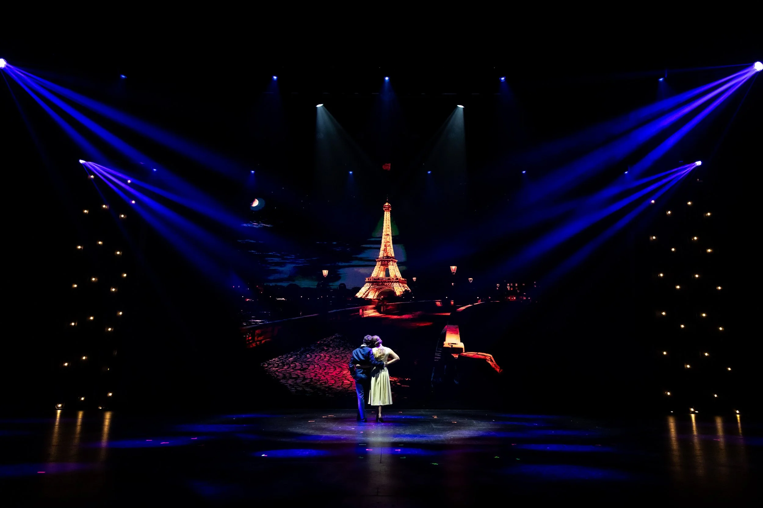 Stage scene with a couple dancing near the Eiffel Tower, illuminated with colorful lights and spotlights, in a theatrical performance.
