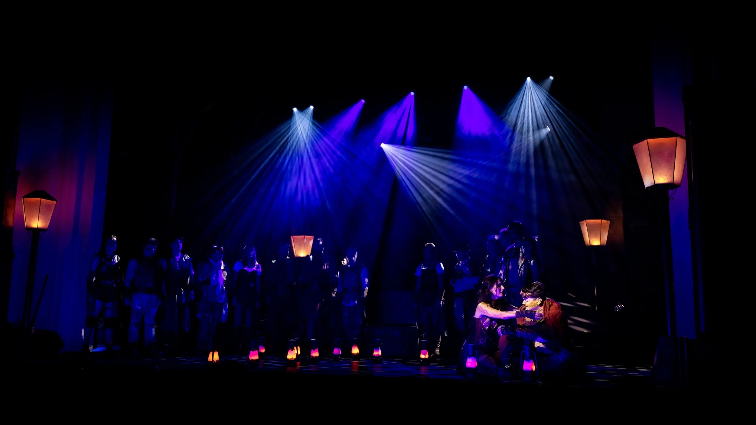 Stage scene with performers and crew under purple and blue lighting, with some seated at a table and others standing, surrounded by lanterns on the floor, in a dark theater environment.