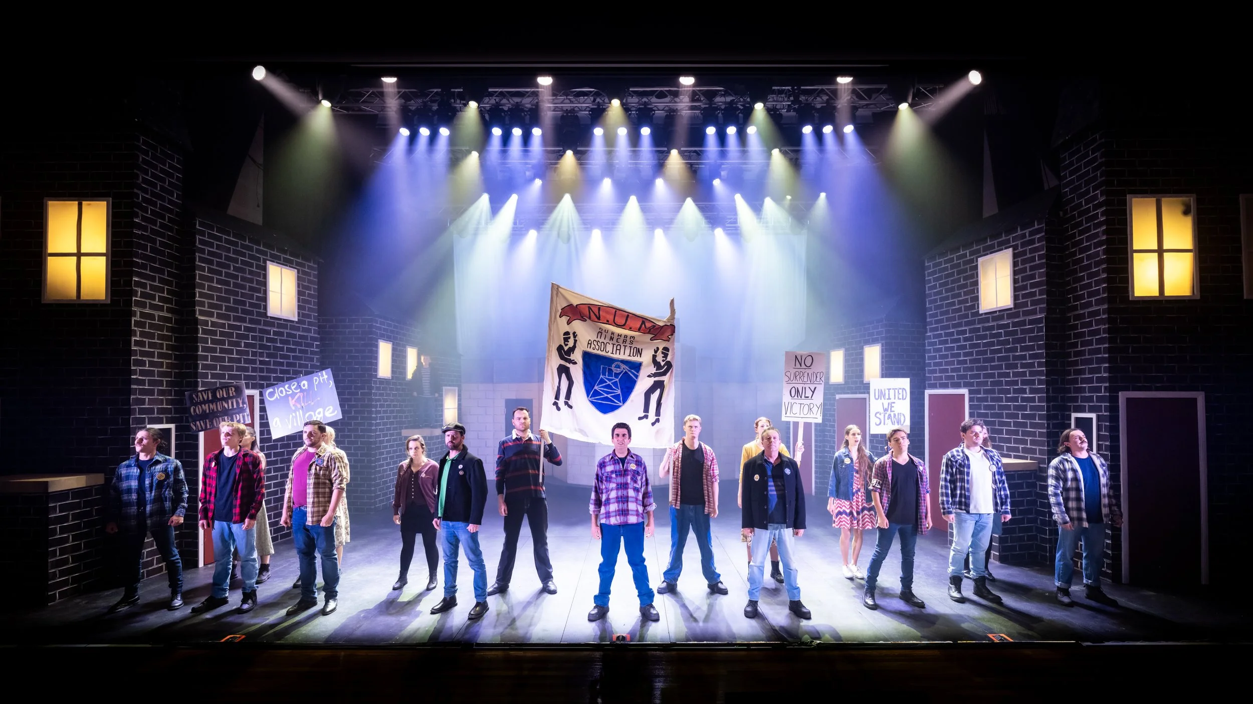 On a theater stage, a group of people are standing with protest signs and a banner. The set is designed to look like a city street with brick buildings and lit windows. The signs include messages like "Save Our Community," "Close a pit, a village," a
