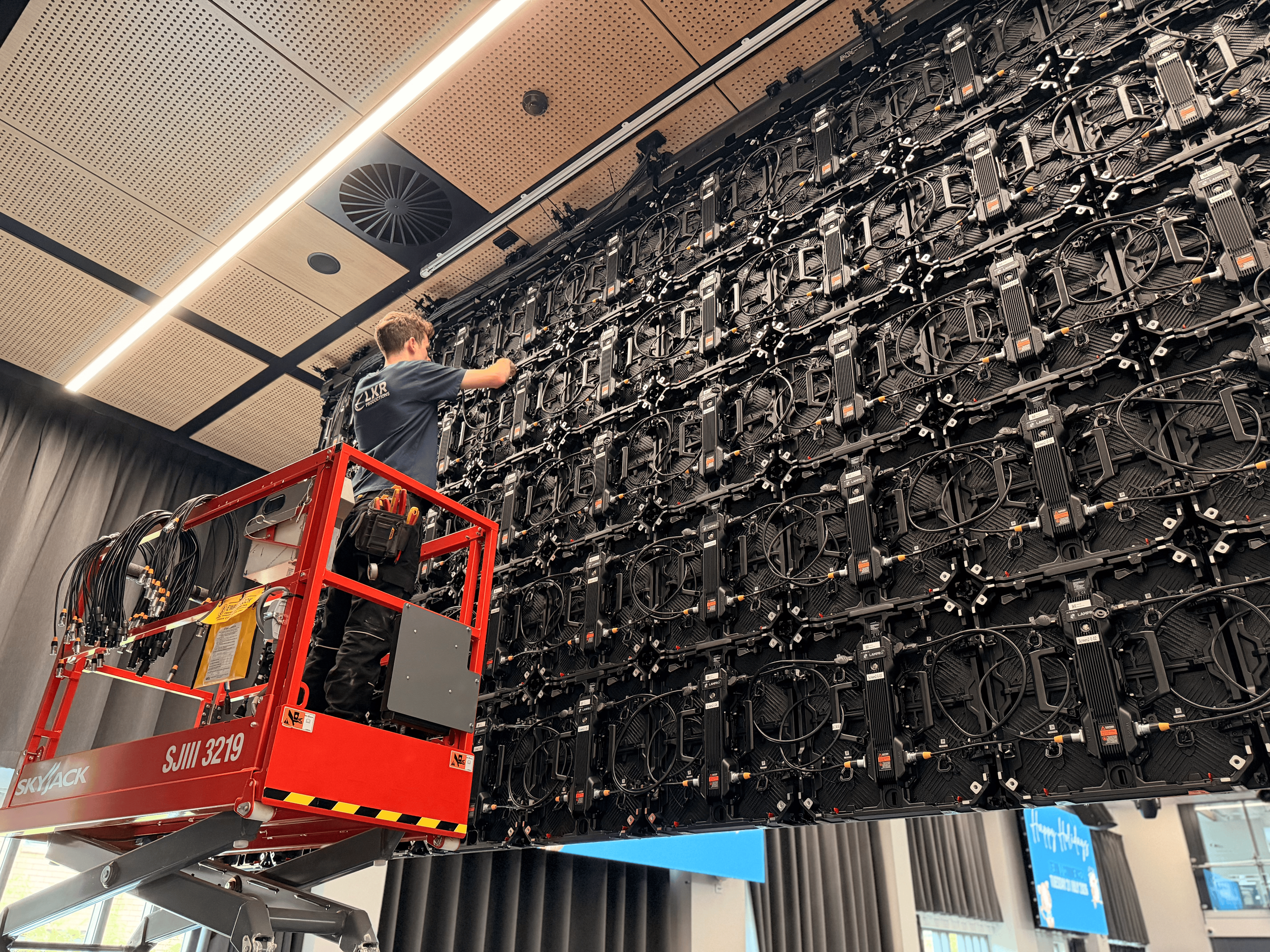 A technician working on a large outdoor LED display board at a commercial location. The technician is on a cherry picker adjusting or installing LED panels.