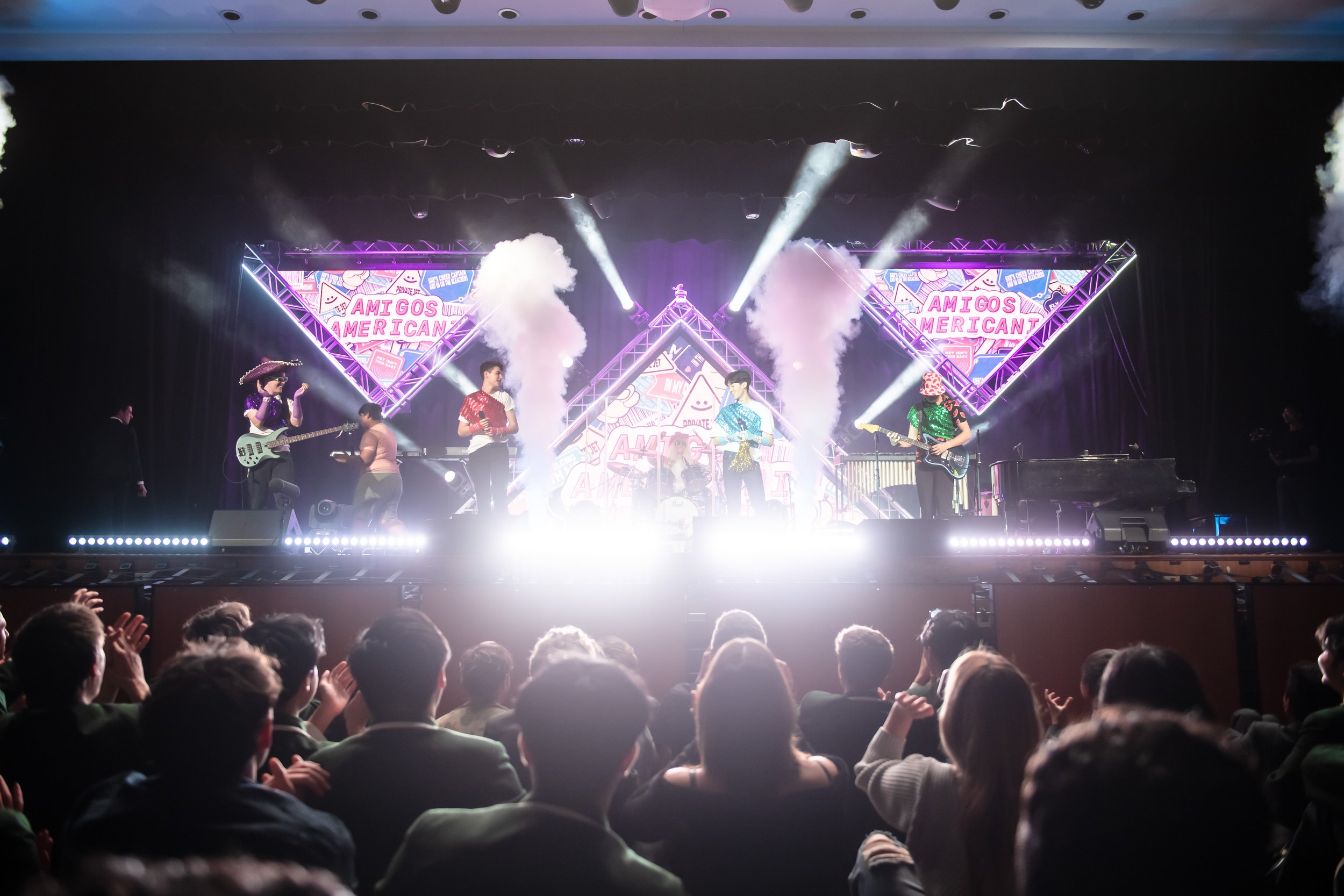 Live performance on stage with band members dressed in colorful costumes, playing guitars and singing, with a vibrant backdrop displaying the words "Amigos Americanz" and animated graphics, audience applauding in the foreground.