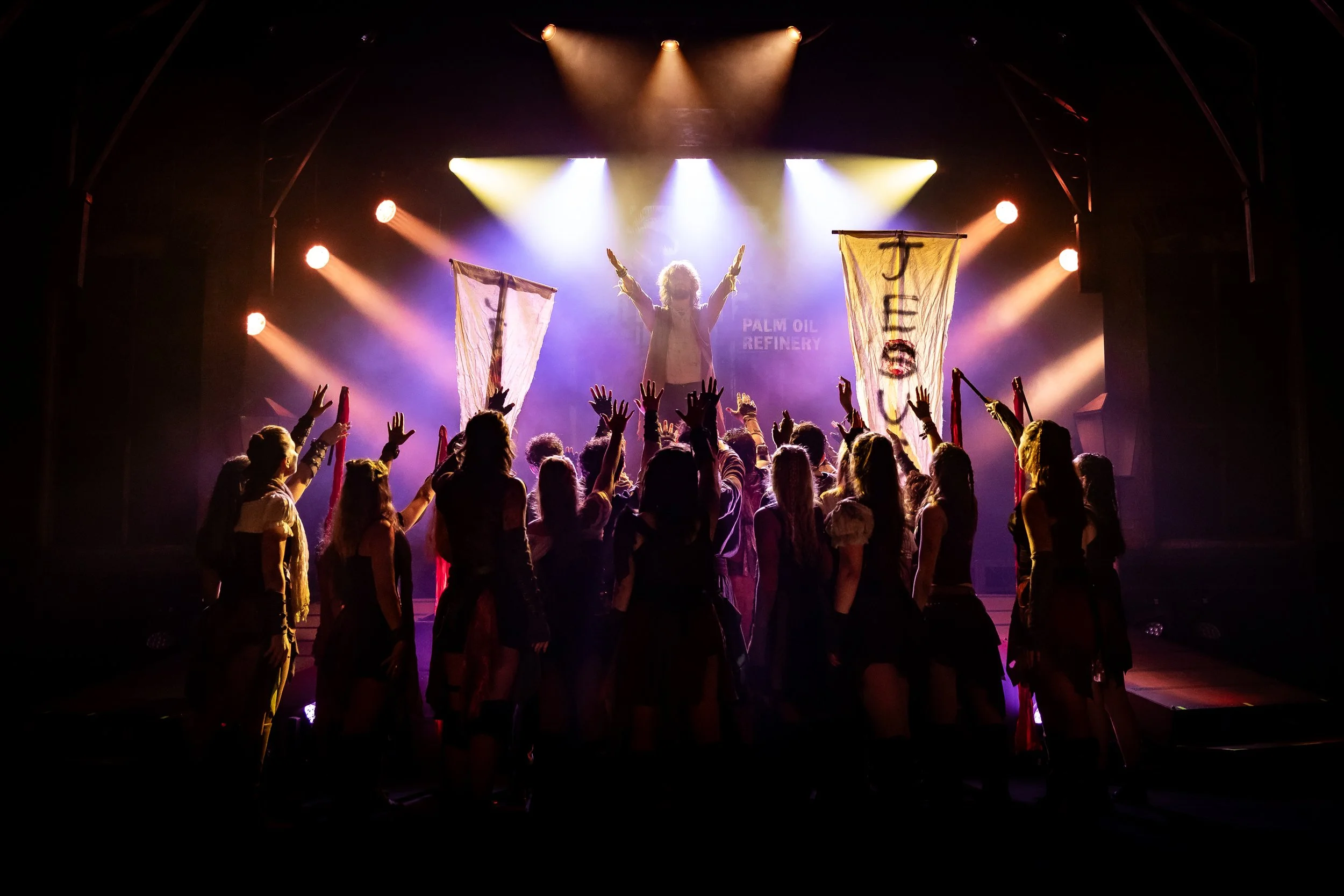 A live concert scene with a singer on stage and a crowd with raised hands in front. The stage is illuminated with colorful lights, and the singer is holding two banners with inscriptions, surrounded by people enjoying the performance.