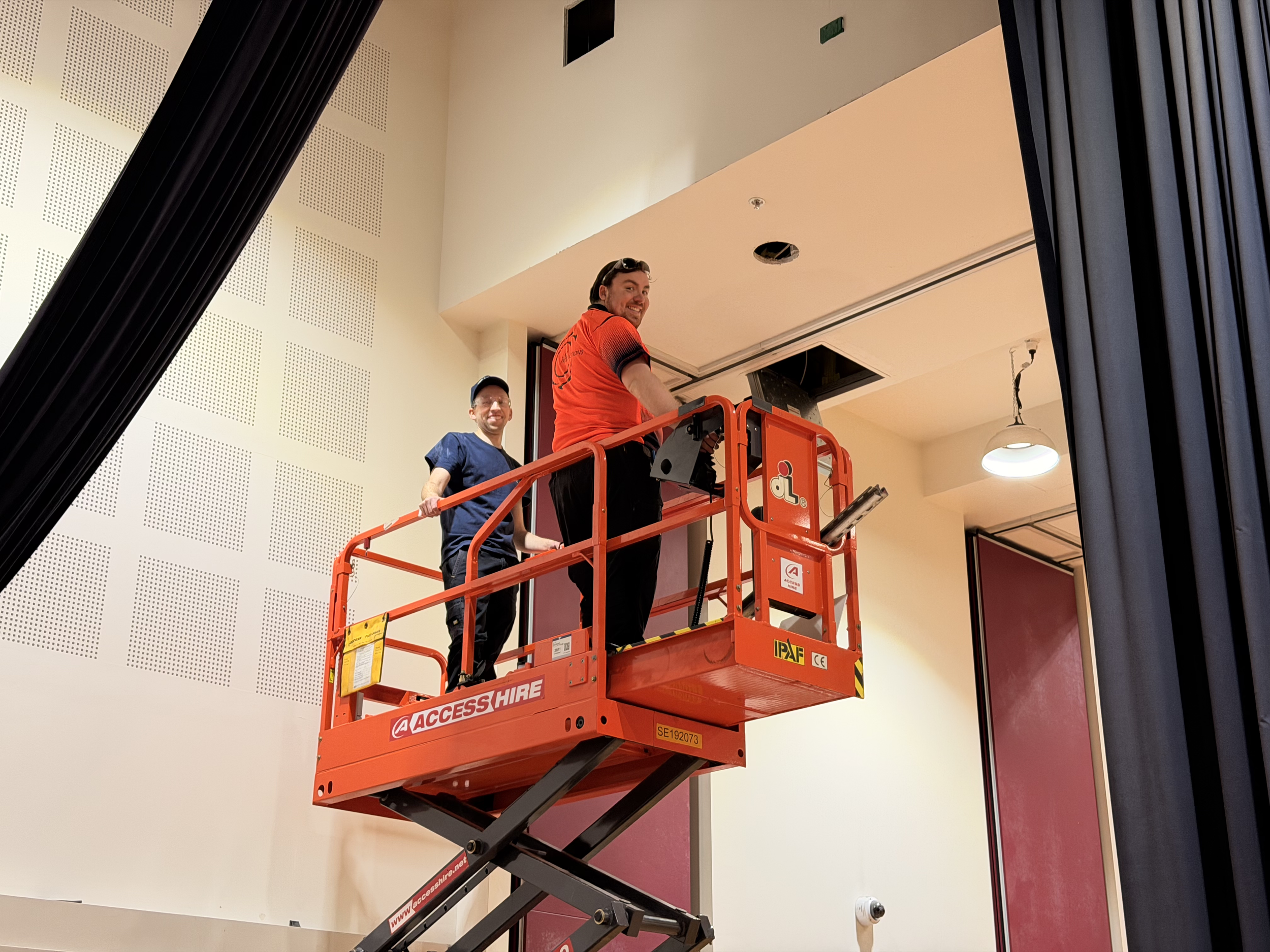 Two workers on a scissor lift installing or repairing a ceiling light in a room with beige walls and dark curtains.