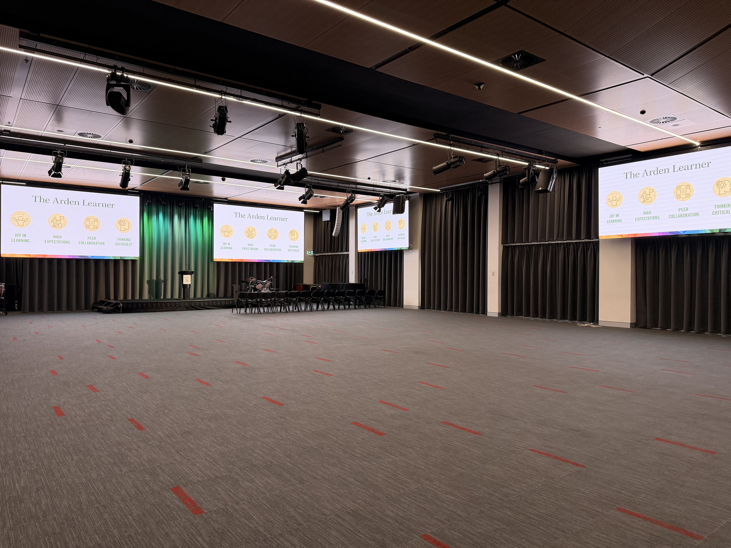 An empty auditorium or conference room with a stage, multiple screens displaying a presentation titled "The Arden Learner," and rows of chairs. The room has a ceiling with linear lighting, black curtains at the sides of the stage, and various audio and lighting equipment mounted on the ceiling.