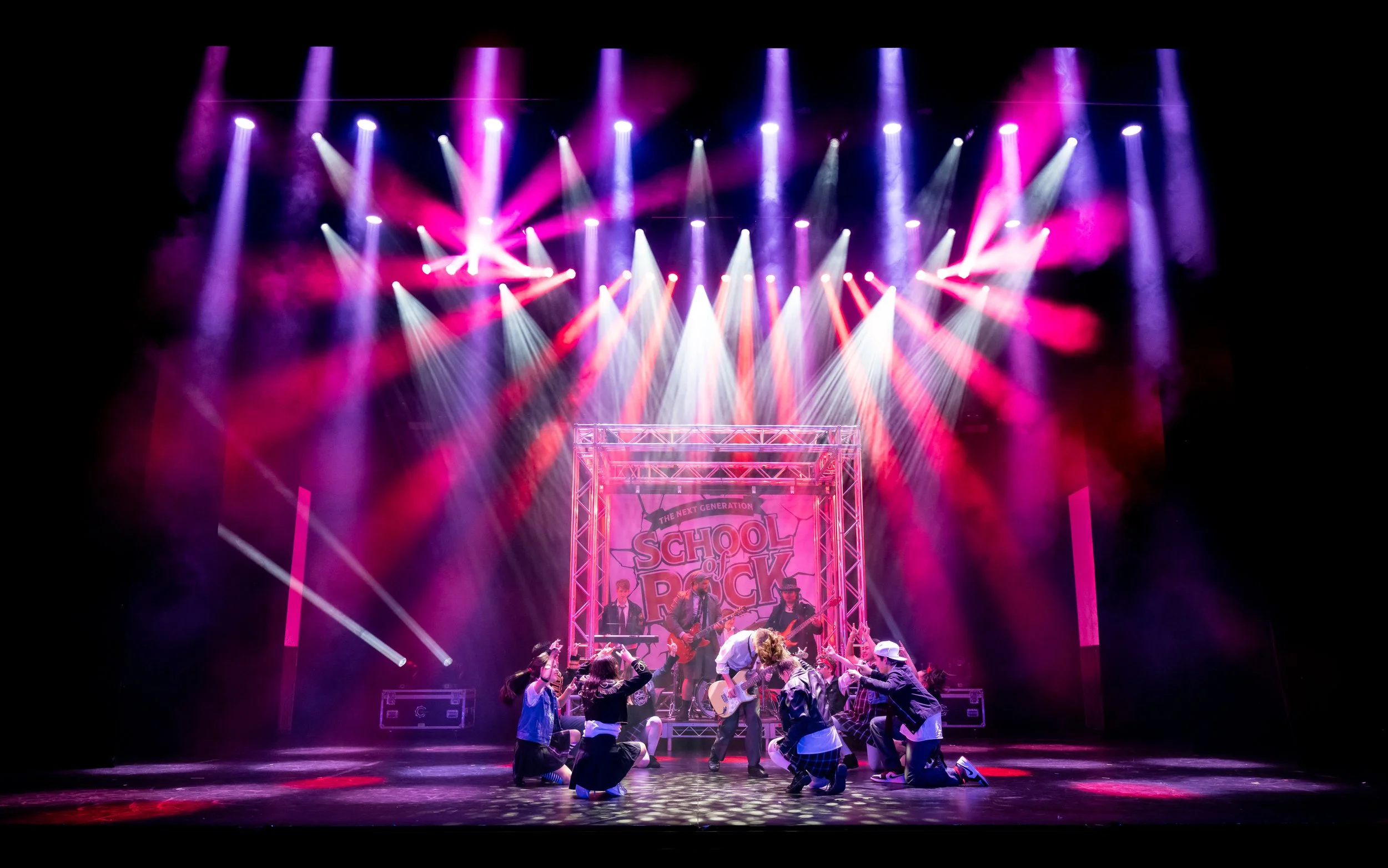 Stage with colorful lights and a band performing at a school of rock event, with children dancing in front.