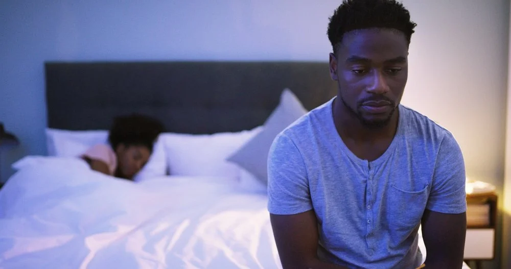man sitting on the edge of his bed looking upset while his wife is sleeping