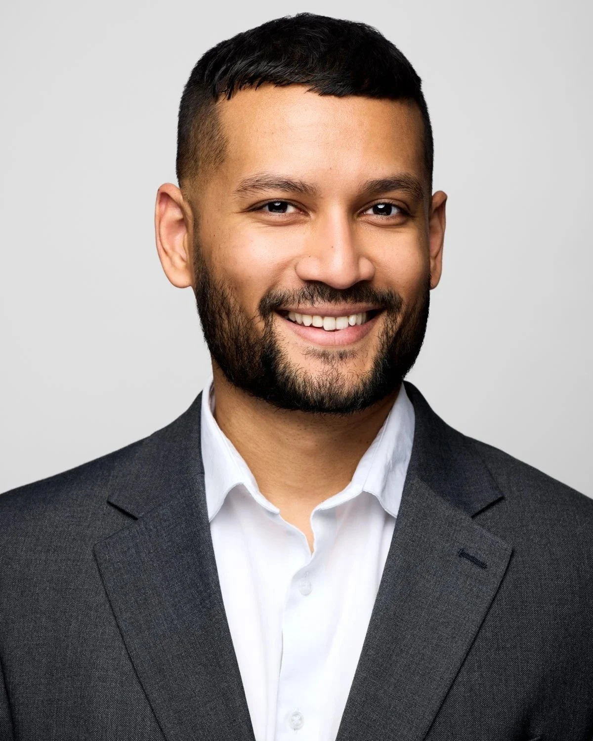 A professional corporate headshot of a smiling man in a grey blazer, shot against a clean white background in a Toronto studio.