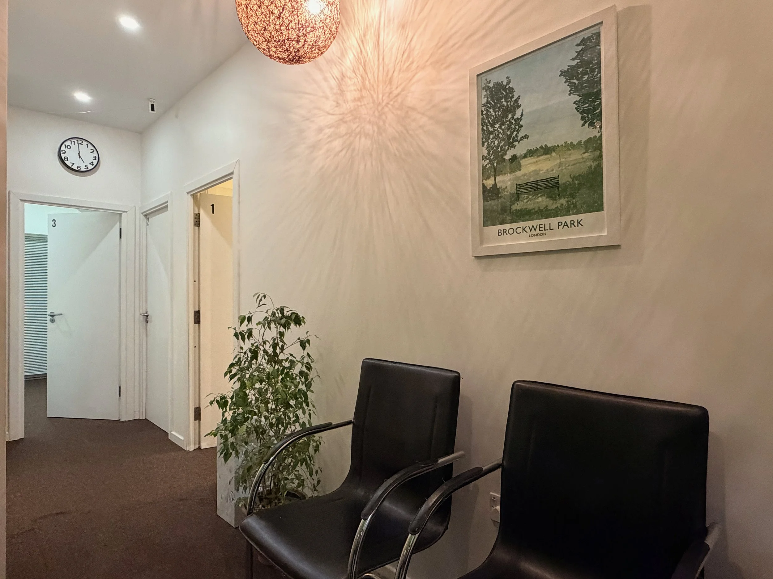 Waiting area with two black chairs, a potted plant, a framed picture of Brockwell Park, a clock on the wall, and three doors numbered 1, 2, and 3.