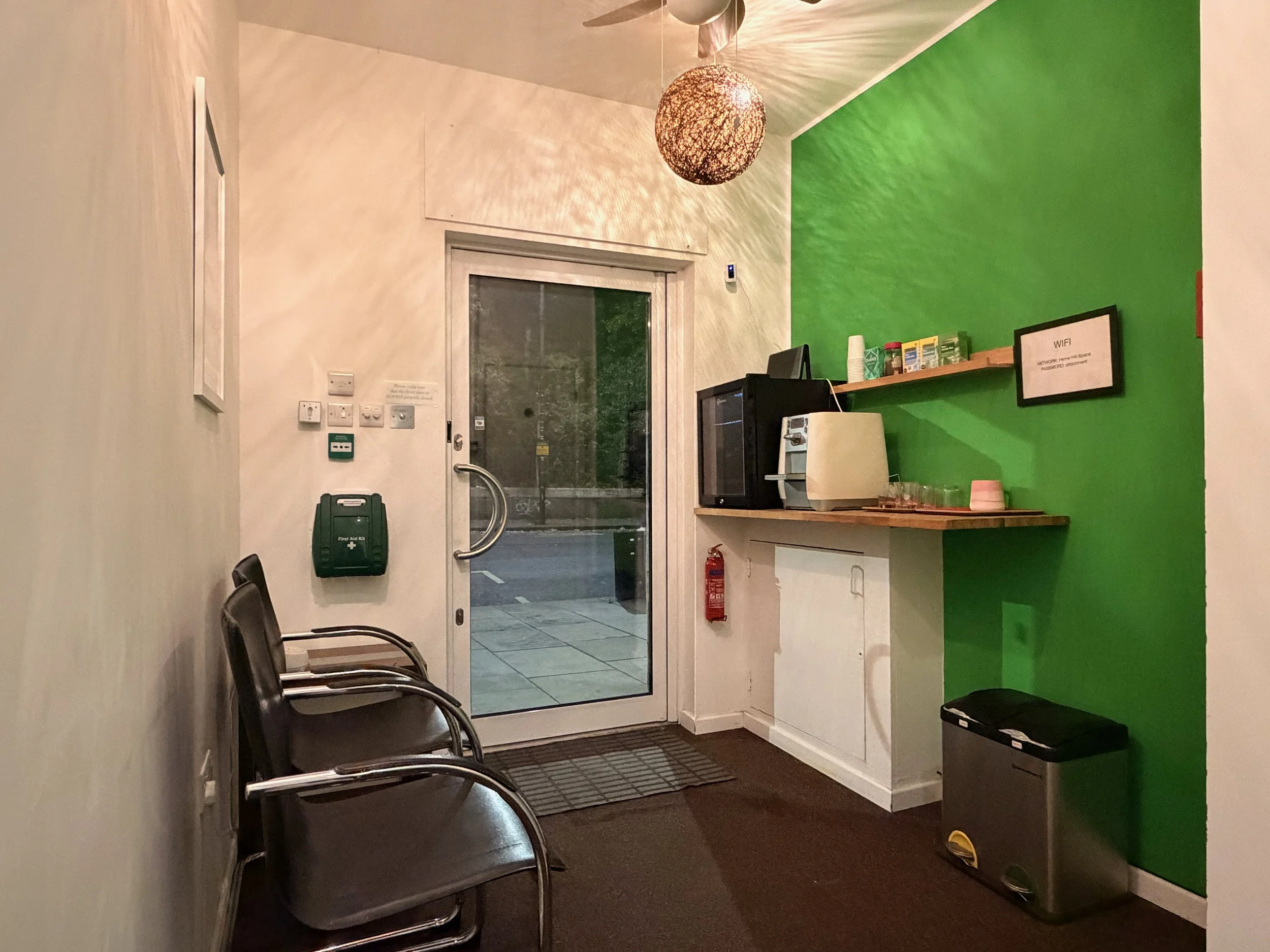 Interior of a small lobby with a green accent wall, a glass door, chairs, a clipboard, a fire alarm, a mounted first aid kit, a small fridge, a coffee machine, shelves with supplies, and a small trash can.