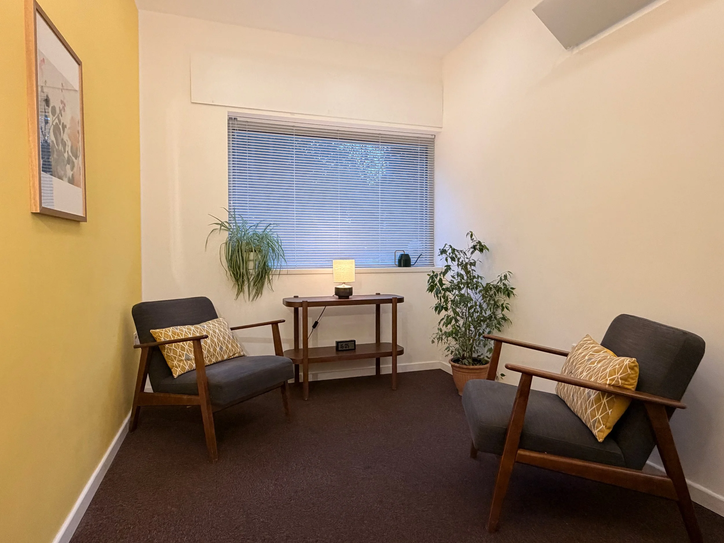 A cozy therapy room with two dark gray armchairs with wooden frames and yellow patterned pillows, a small wooden side table with a lamp, a large window with blinds, and two potted plants with a yellow accent wall on the left and a framed artwork.