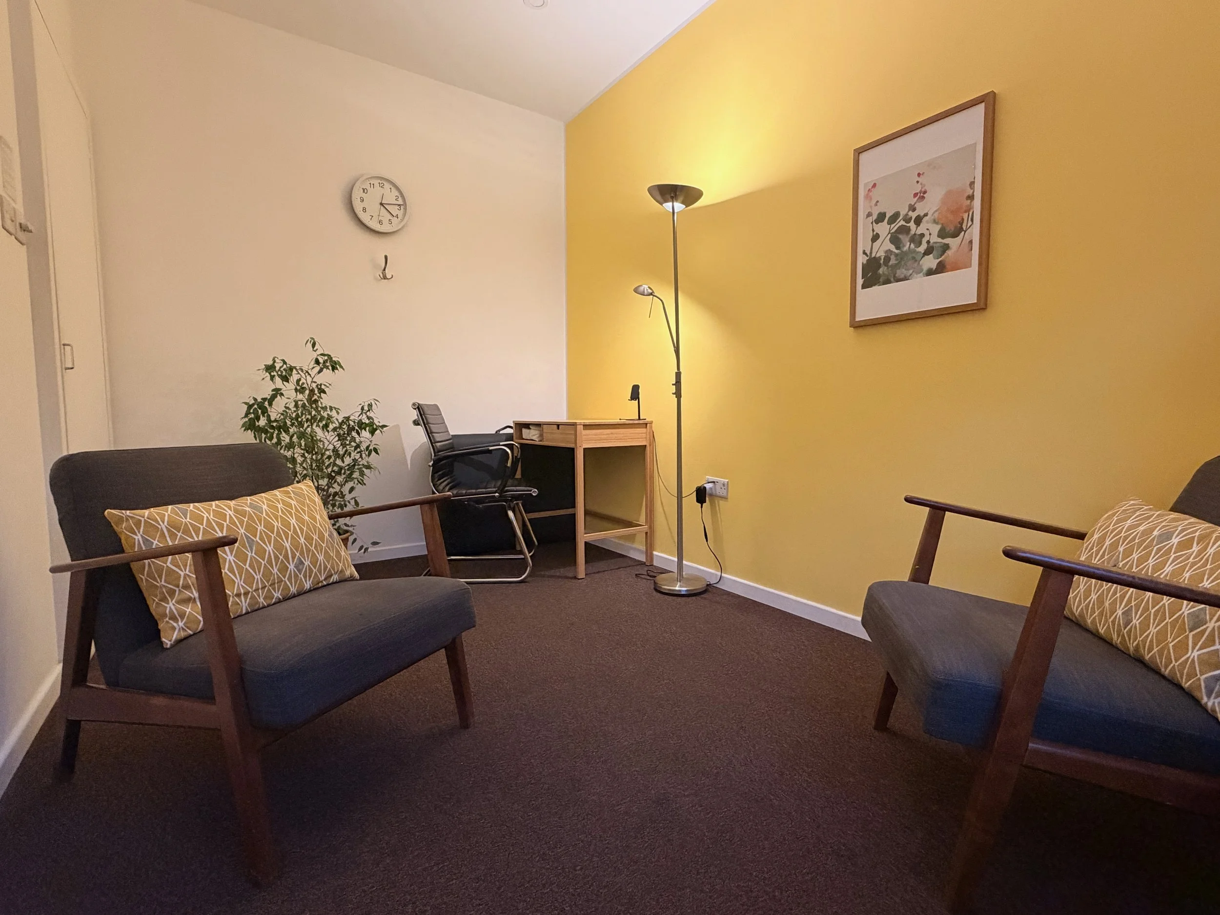 A cozy therapy room with two gray armchairs with wooden frames and yellow patterned throw pillows, a black office chair, a small wooden desk, a tall silver floor lamp, a framed floral artwork on a yellow wall, a wall clock, a potted plant.