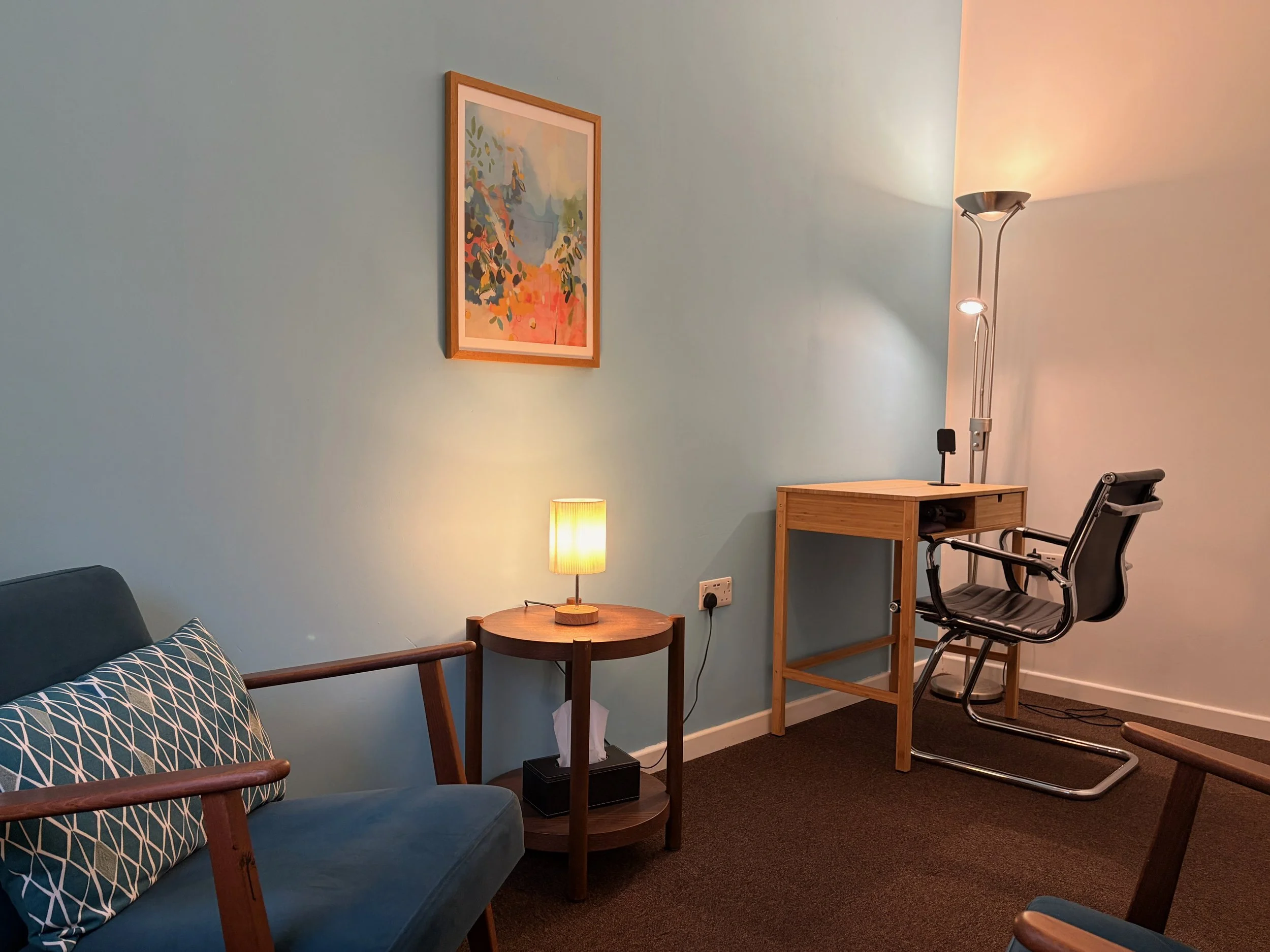 A cozy therapy room with a blue sofa and geometric-patterned pillow, a small wooden side table with a lamp, a wooden desk with a black office chair, a tall floor lamp, and a framed abstract painting on the wall.