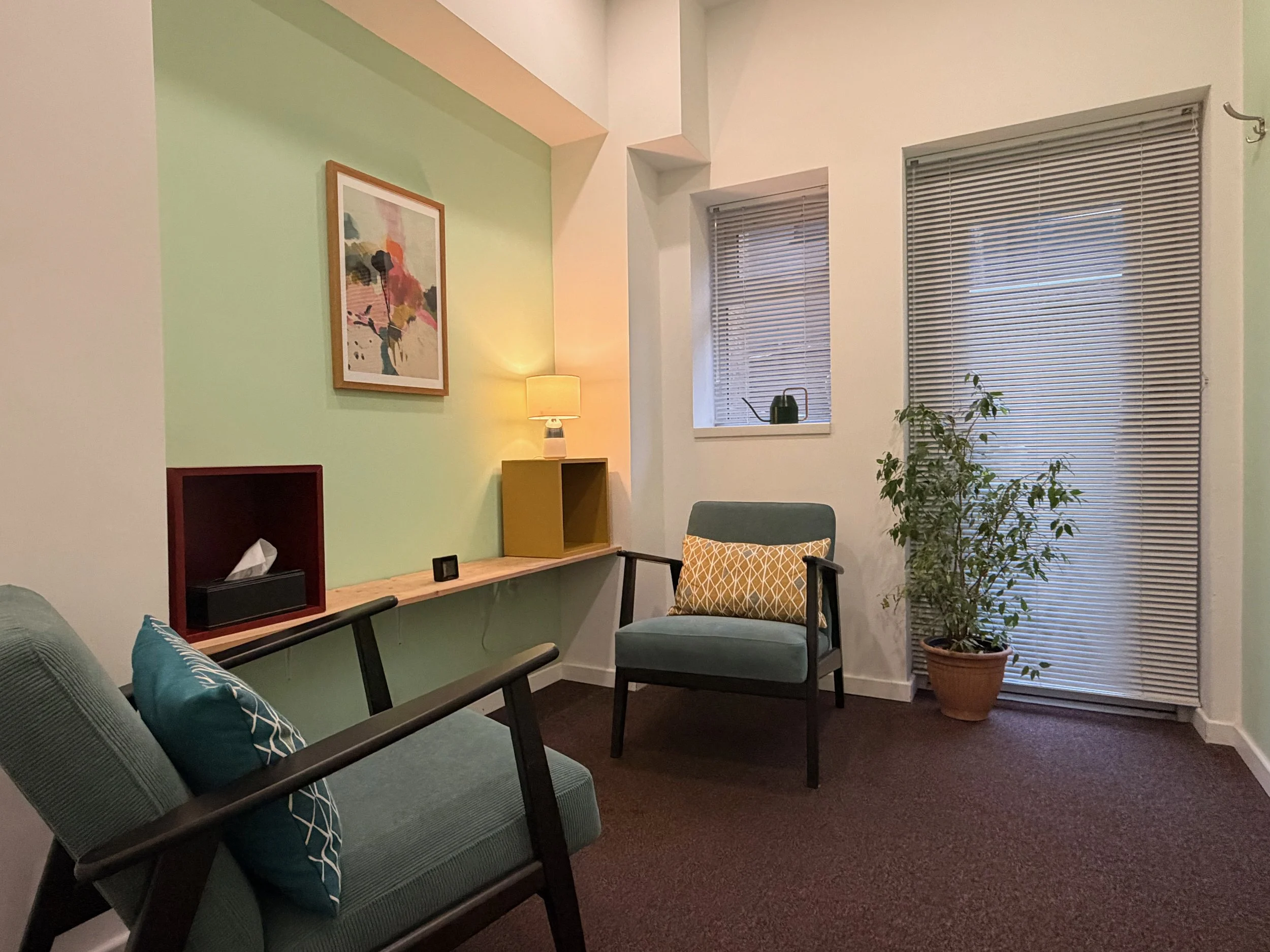 A cozy therapy room with pastel green accent wall, a framed abstract painting, a small table lamp, and two chairs with decorative pillows. There is a potted plant and window blinds on the windows.
