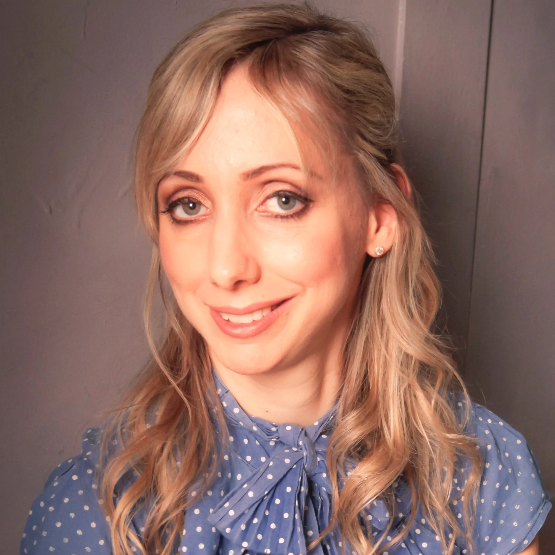 A woman with blonde wavy hair and blue eyes, smiling, wearing a blue polka dot blouse with a bow tie, standing against a gray background.