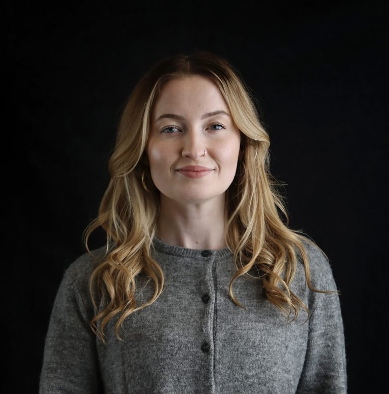 A woman with blonde hair, wearing a grey sweater, standing against a black background.
