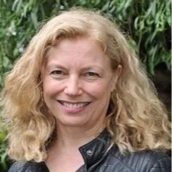 Portrait of a woman with curly blonde hair smiling, wearing a leather jacket outdoors.