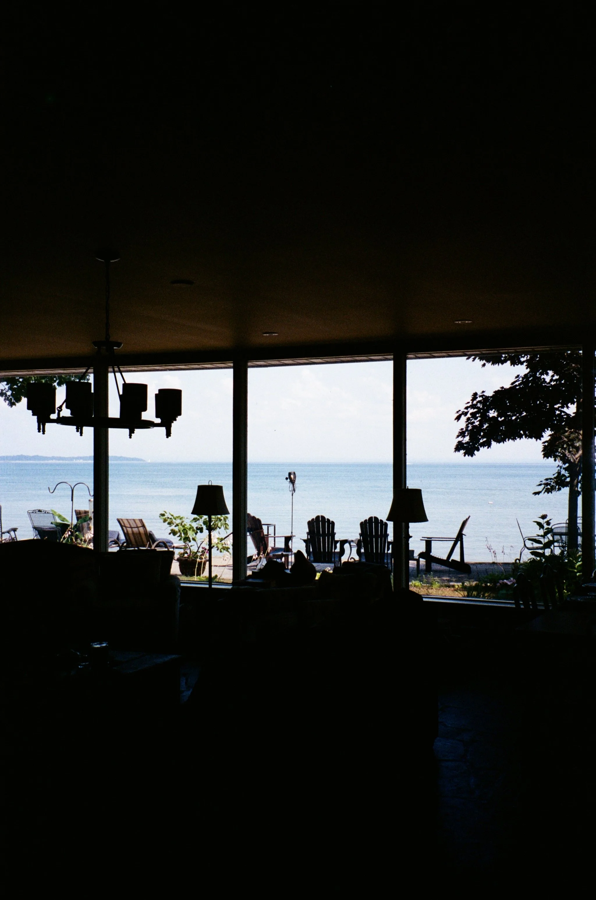 View of a waterfront patio with outdoor furniture, overlooking the ocean, under a covered area with a chandelier hanging from the ceiling, and a tree on the right side.