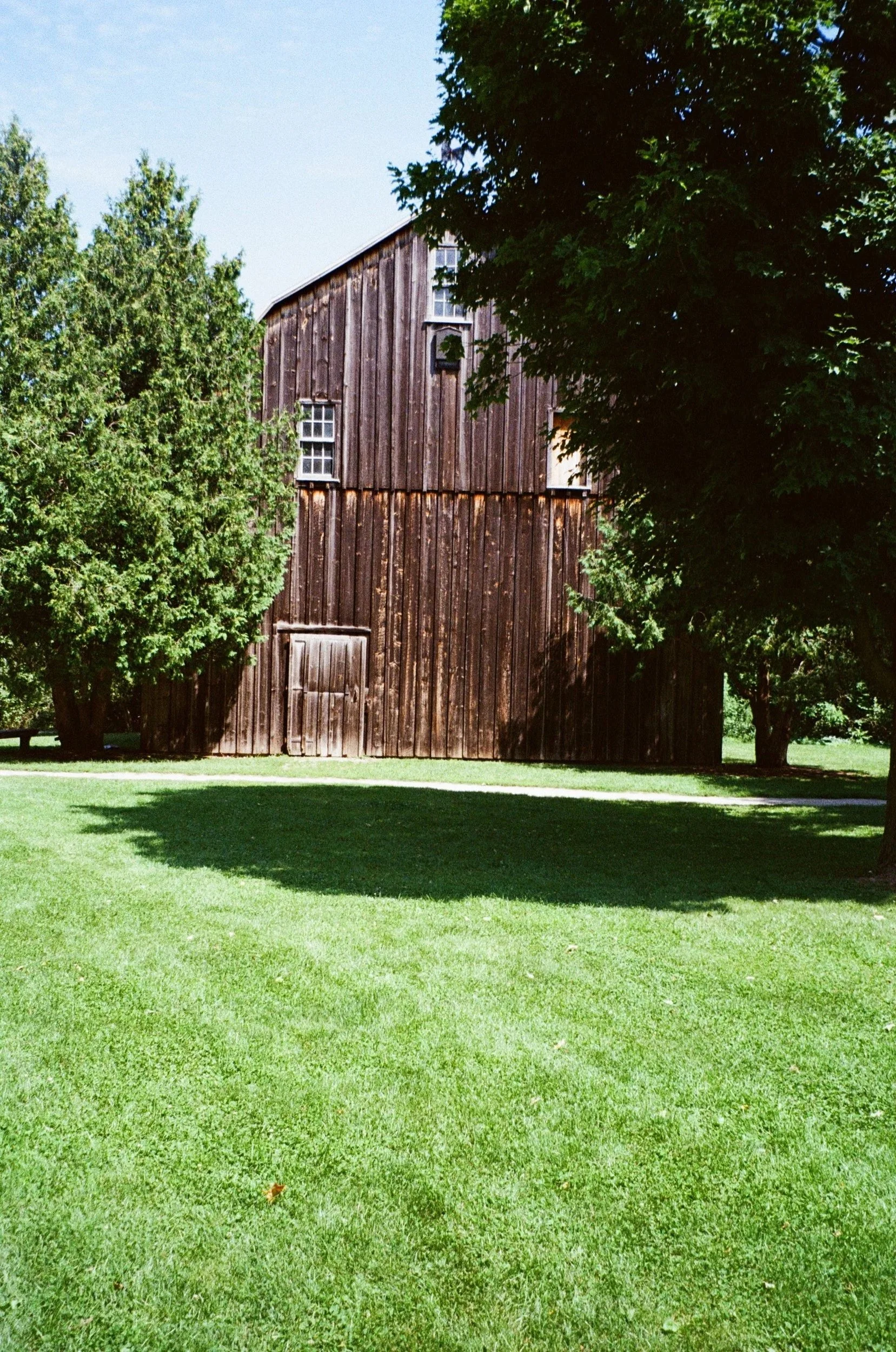 A wooden barn surrounded by green trees and a well-maintained green lawn under a blue sky.