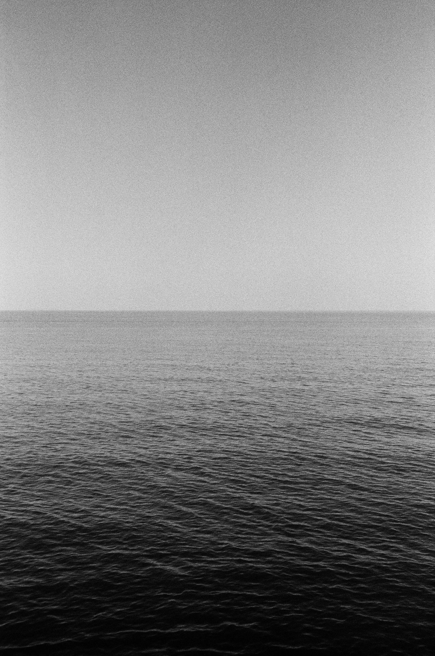 A grayscale photo of a calm ocean with a horizon, showing subtle waves and a gradient sky.