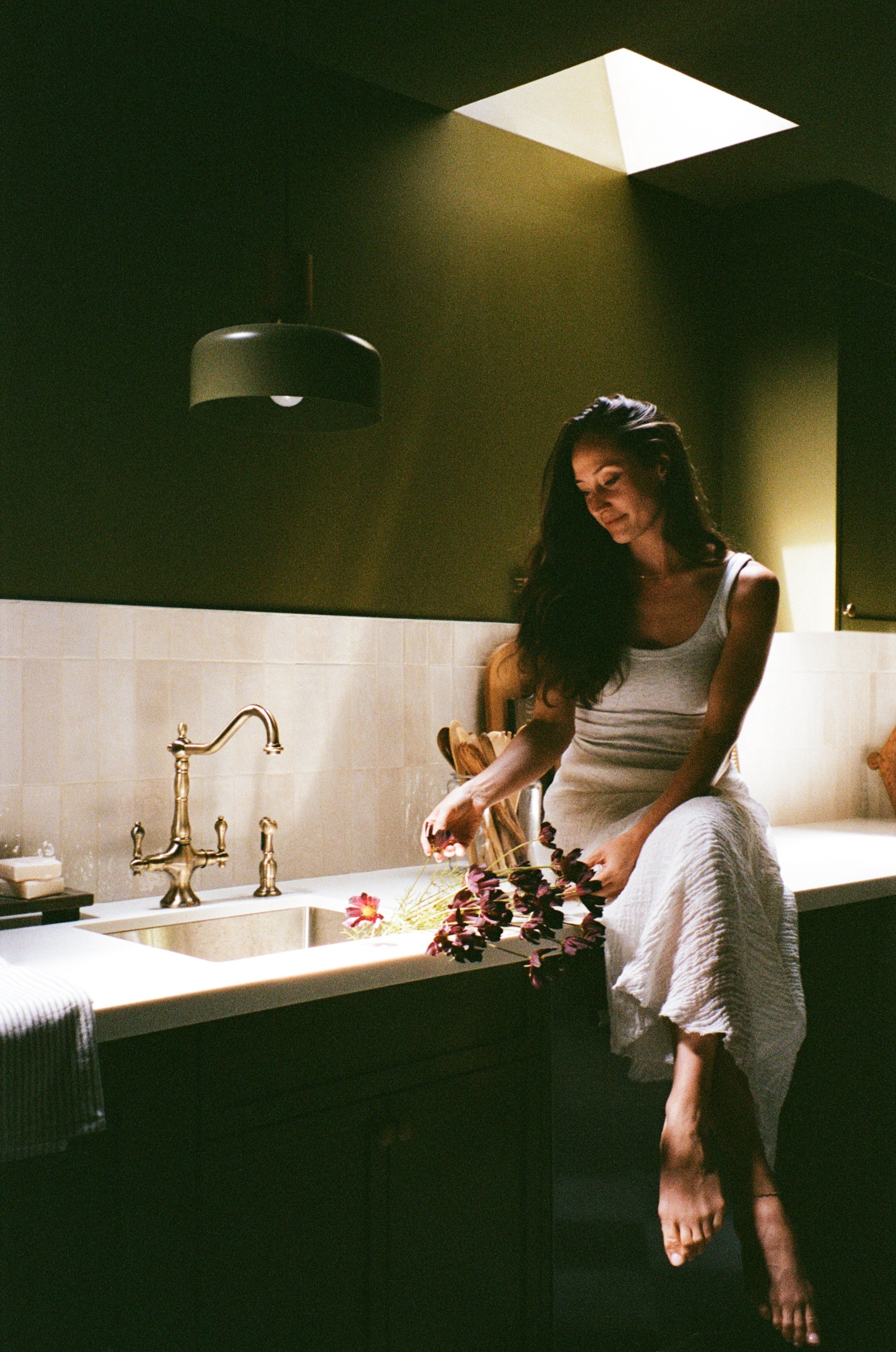 A woman with long dark hair, wearing a white tank top and a white towel around her waist, is arranging flowers on a kitchen countertop under a skylight in a dimly lit room.