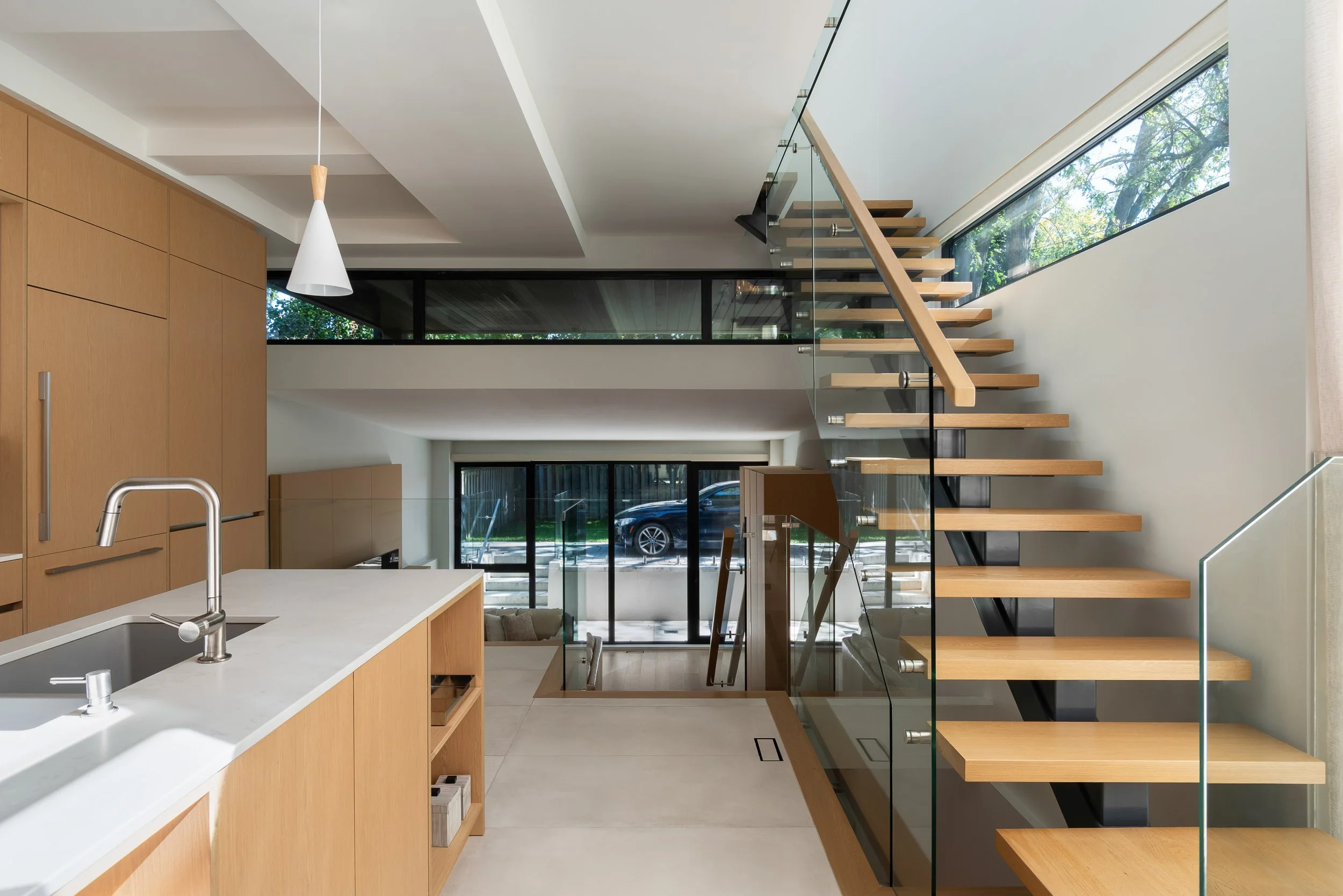 Interior view of a modern two-story home with an open kitchen and staircase, featuring wooden cabinets, a white countertop, glass railing, and large windows showing trees outside.
