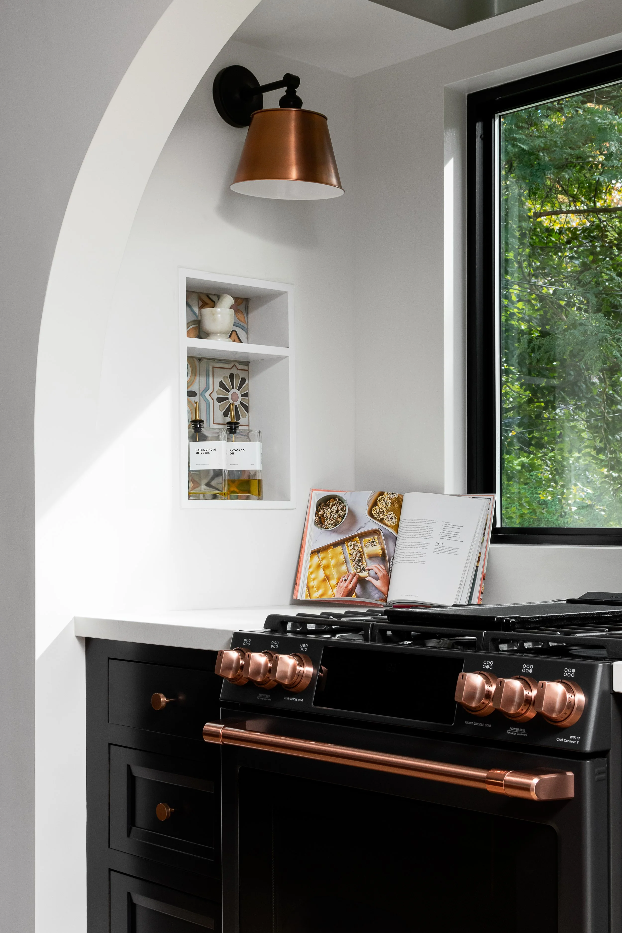 A modern kitchen stove with copper knobs and handle, a white textured wall with built-in shelves containing a ceramic bowl and olive oil bottles, a cookbook open on the counter, a window showing green foliage outside, and a copper wall sconce light.