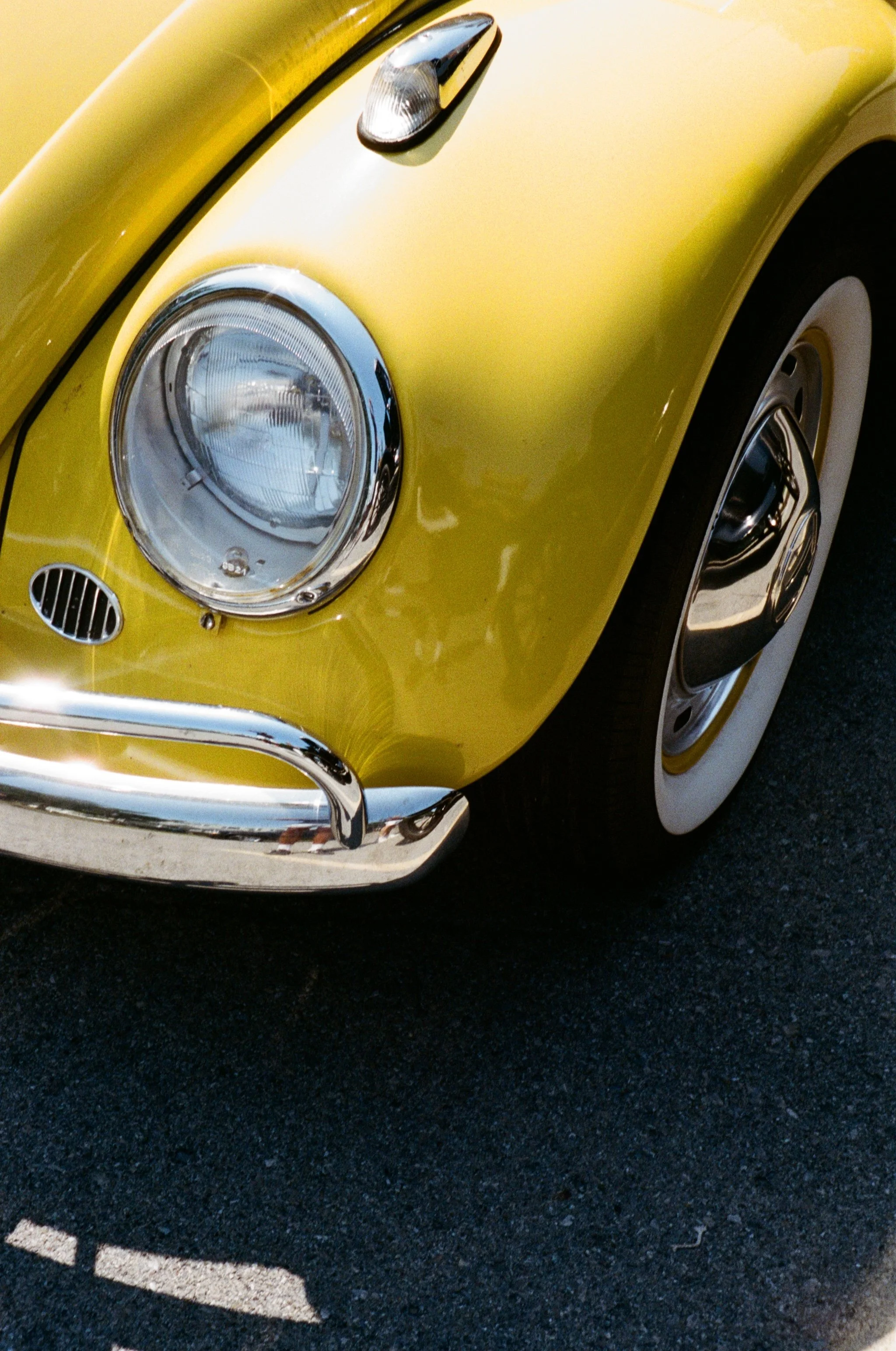 Close-up of a vintage yellow car, showing the front headlight, part of the bumper, and tire on a dark road.