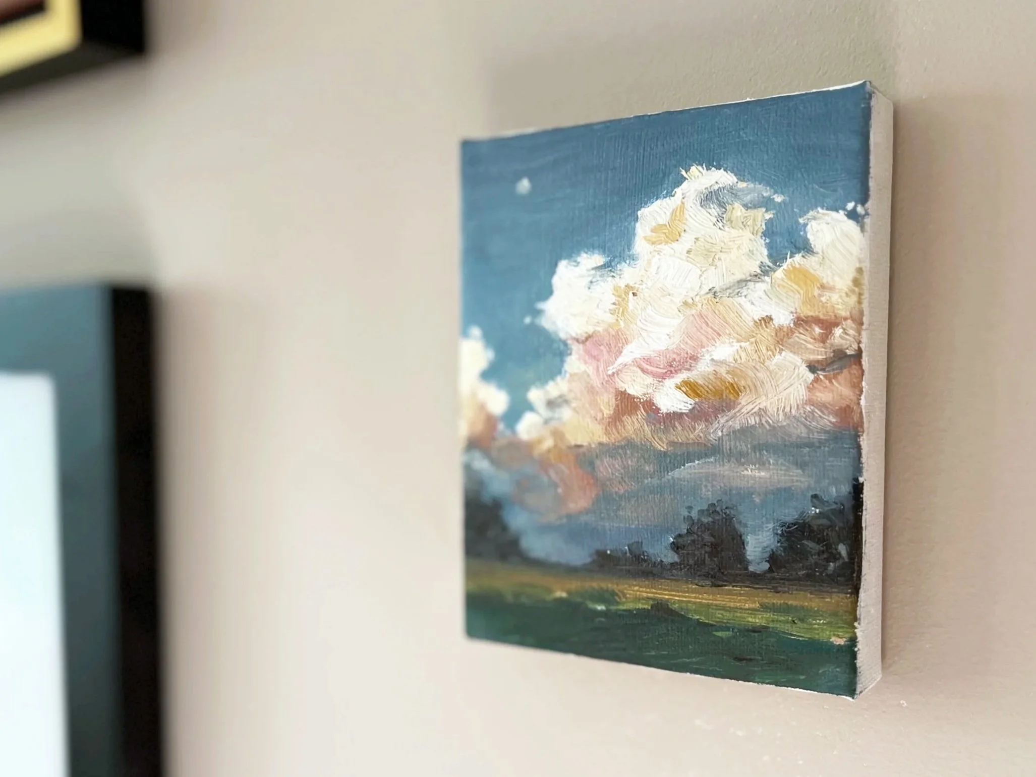A landscape painting on a small canvas depicting a cloud-filled sky with a mix of white, pink, and yellow clouds over a green and brown foresty plain.