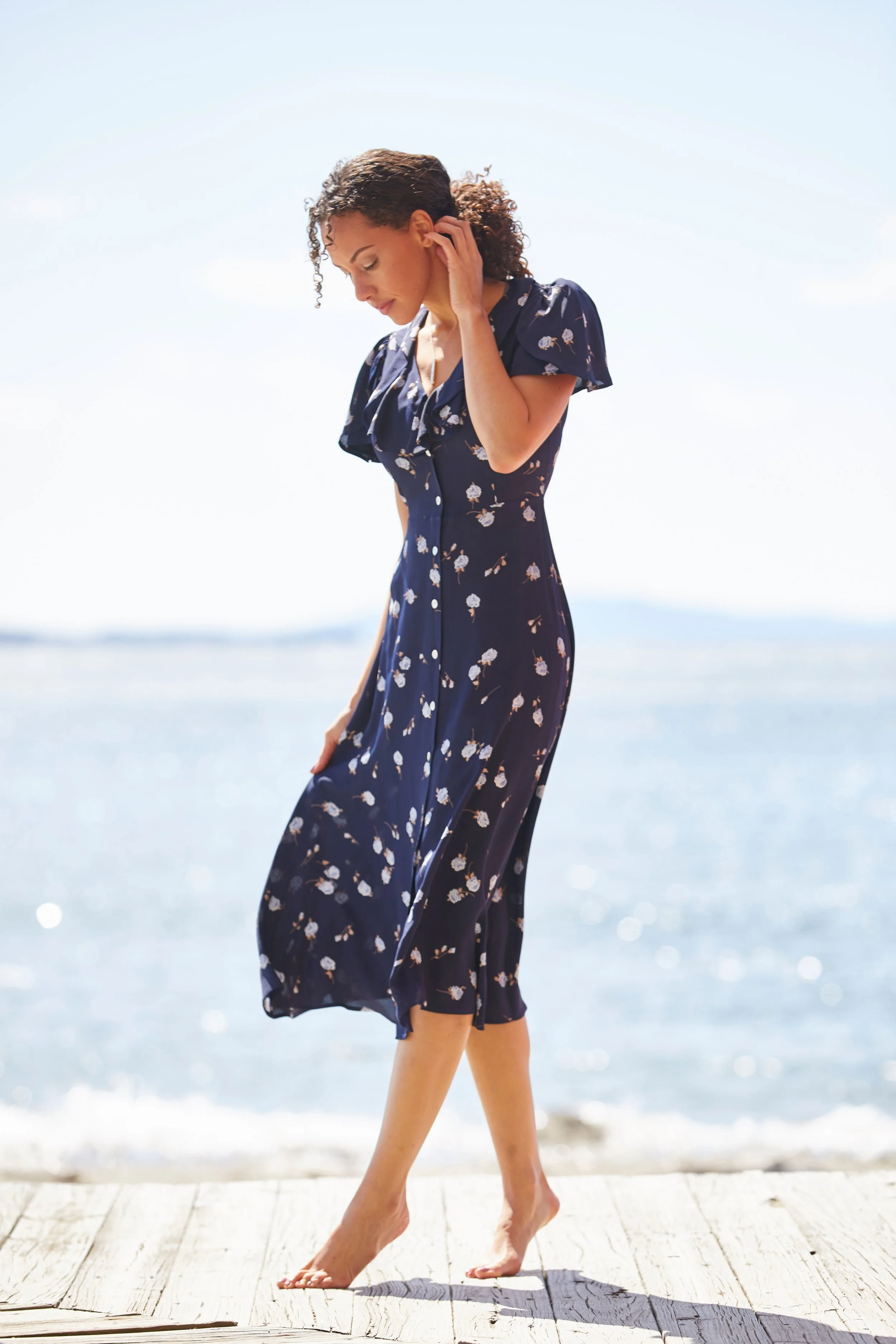 Young woman walking on a boardwalk by the ocean in a blue patterned dress