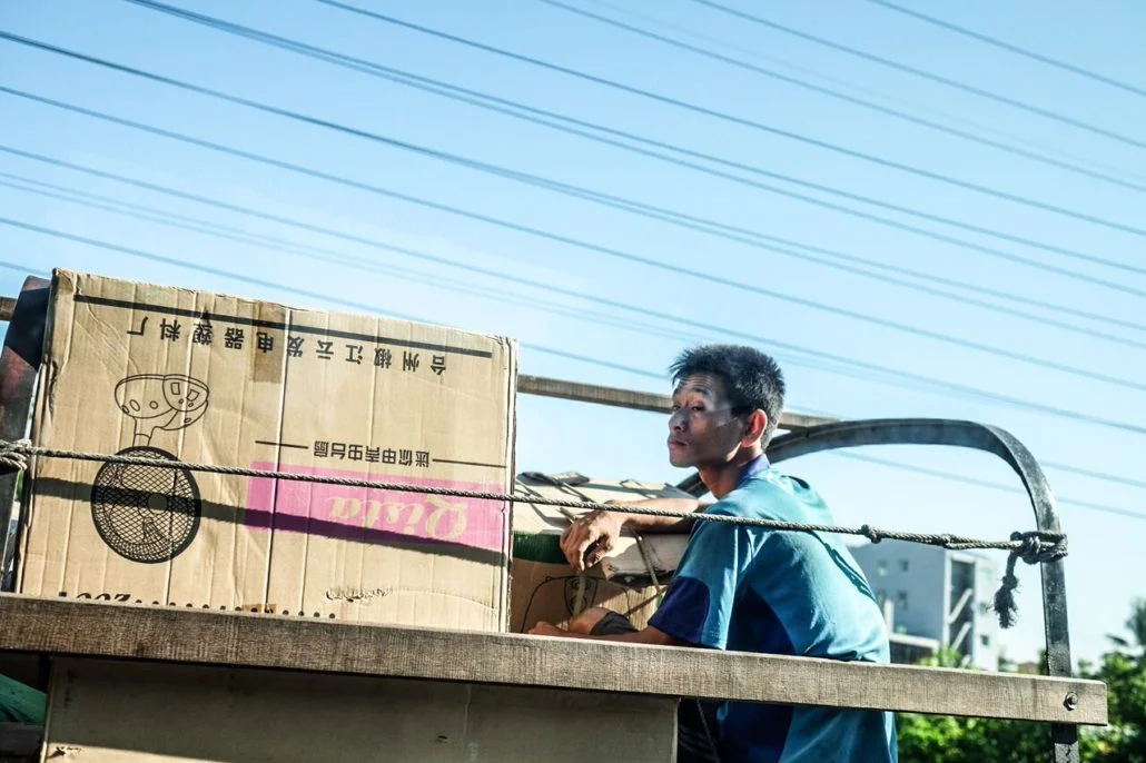 A young man in Myanmar riding on the back of a delivery truck.